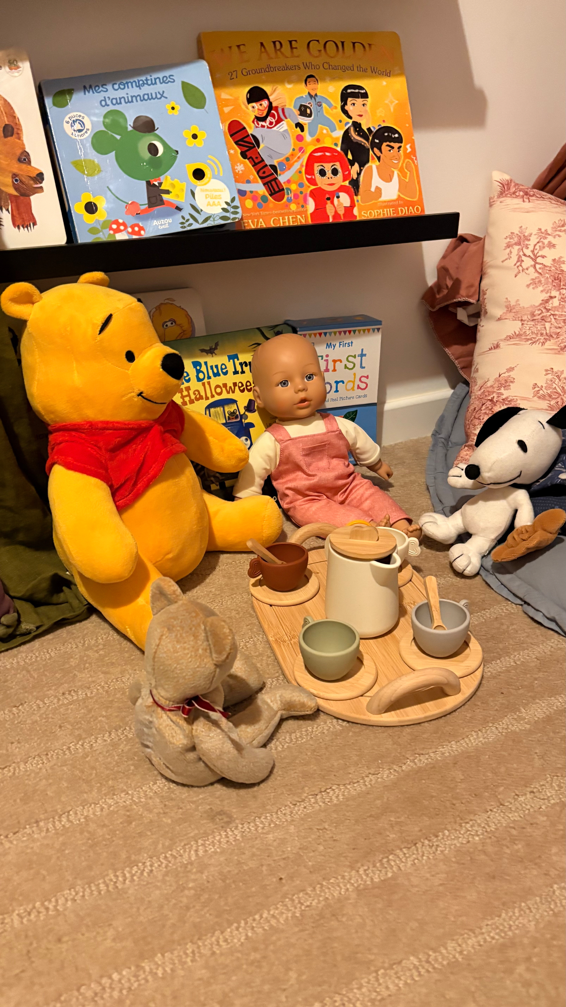 A cozy tea party scene features plush toys and a doll gathered around a wooden tray with cups and a teapot, set against a backdrop of children's books.