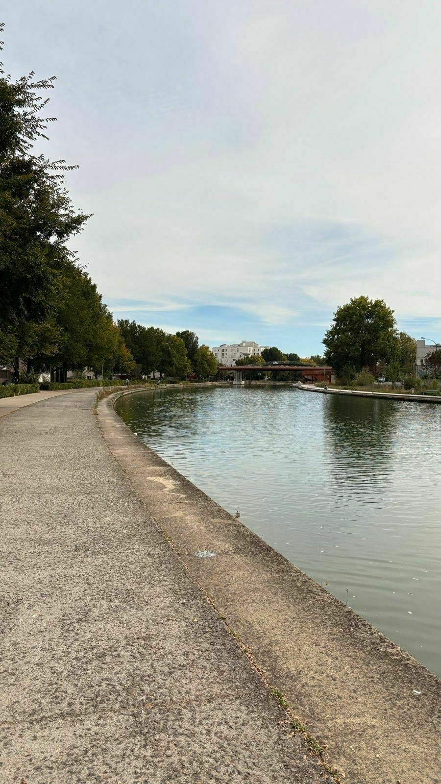 Auto-generated description: A serene canal scene features a walkway lined with trees alongside a gently flowing body of water under a partly cloudy sky.