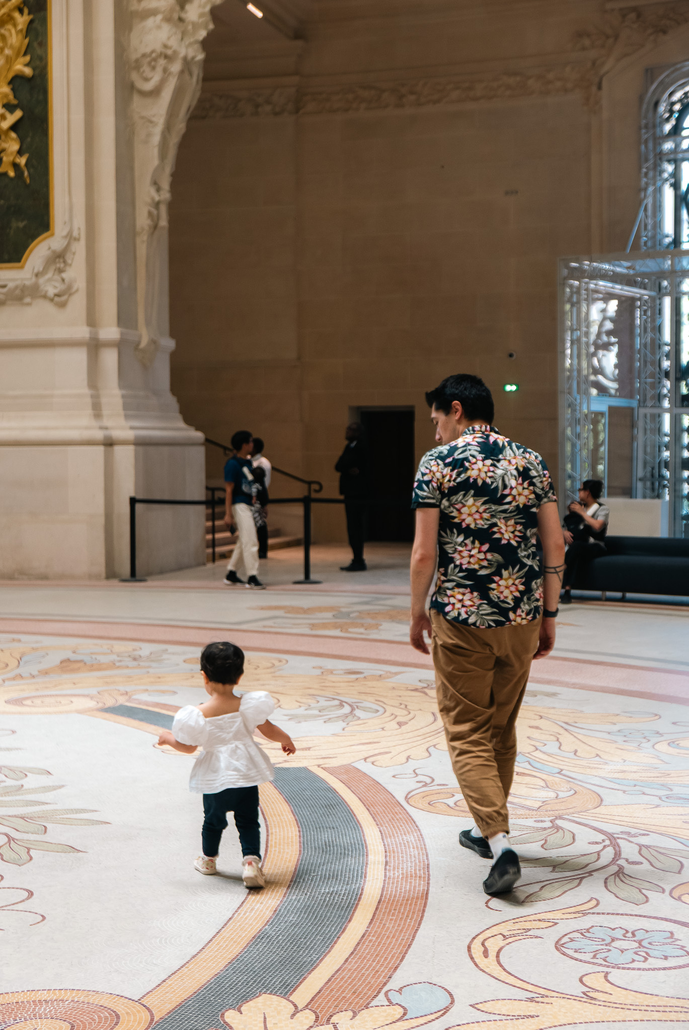 An adult and a child are walking on a decorative floor in an ornate room with large windows.