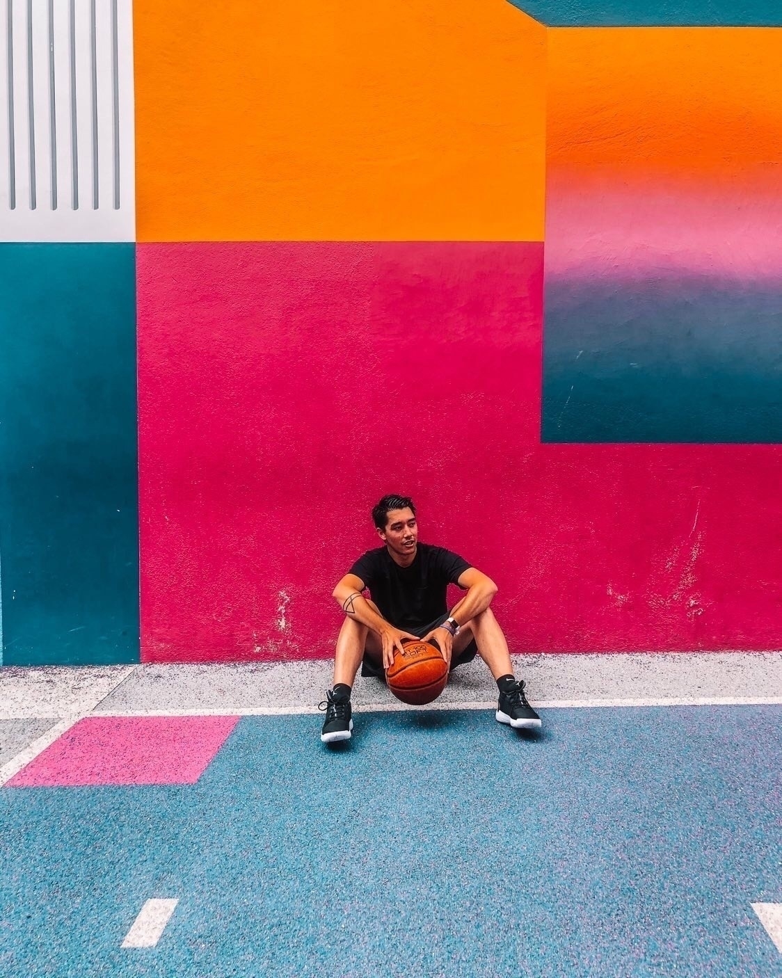 A person sitting on a colorful geometric basketball court, holding a basketball, is set against a vibrant mural background.