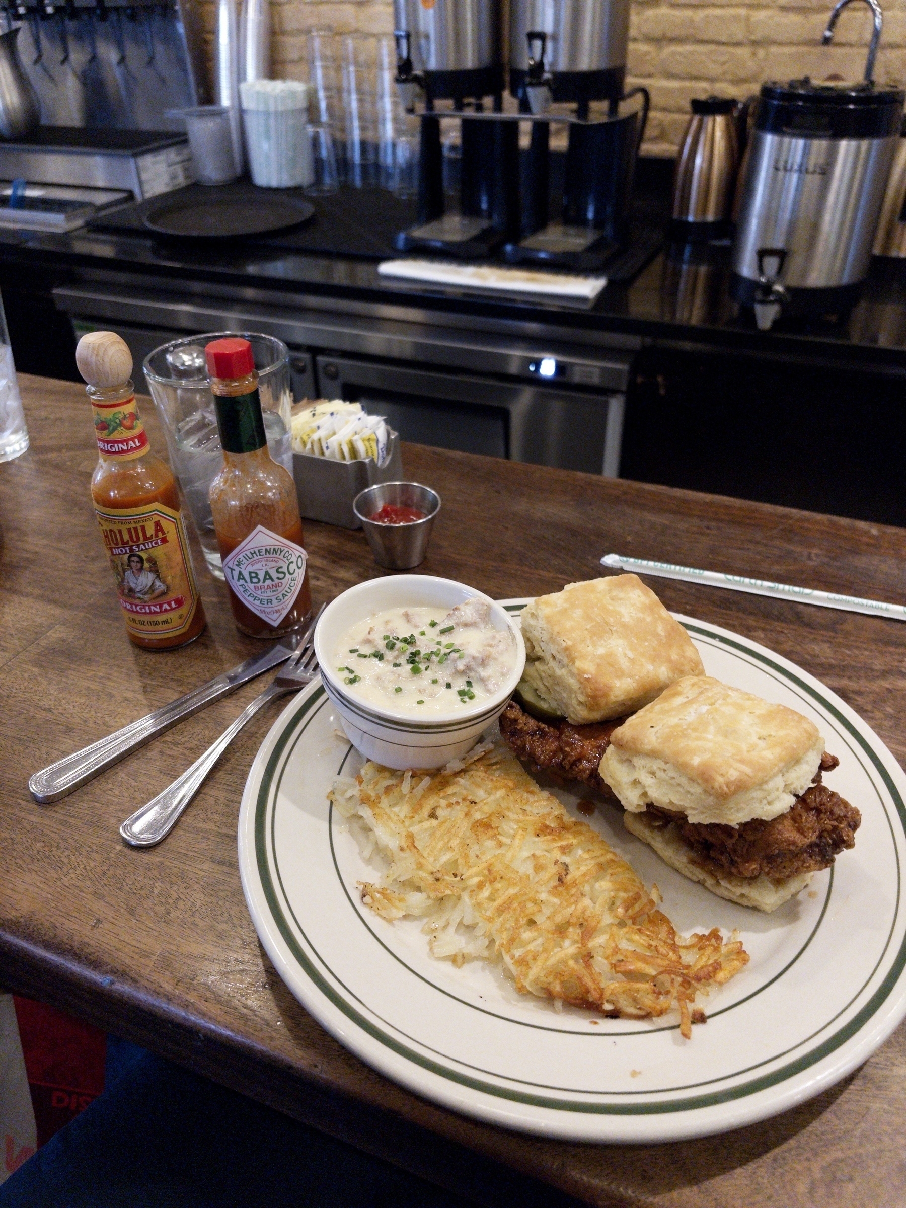 A plate with fried chicken on biscuits, hash browns, and a small bowl of gravy is on a counter next to hot sauce bottles and utensils.