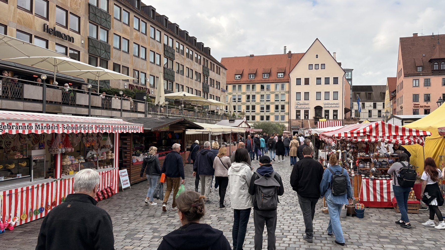 People stroll through a bustling outdoor market with red and white striped stalls, set against a backdrop of traditional European architecture.