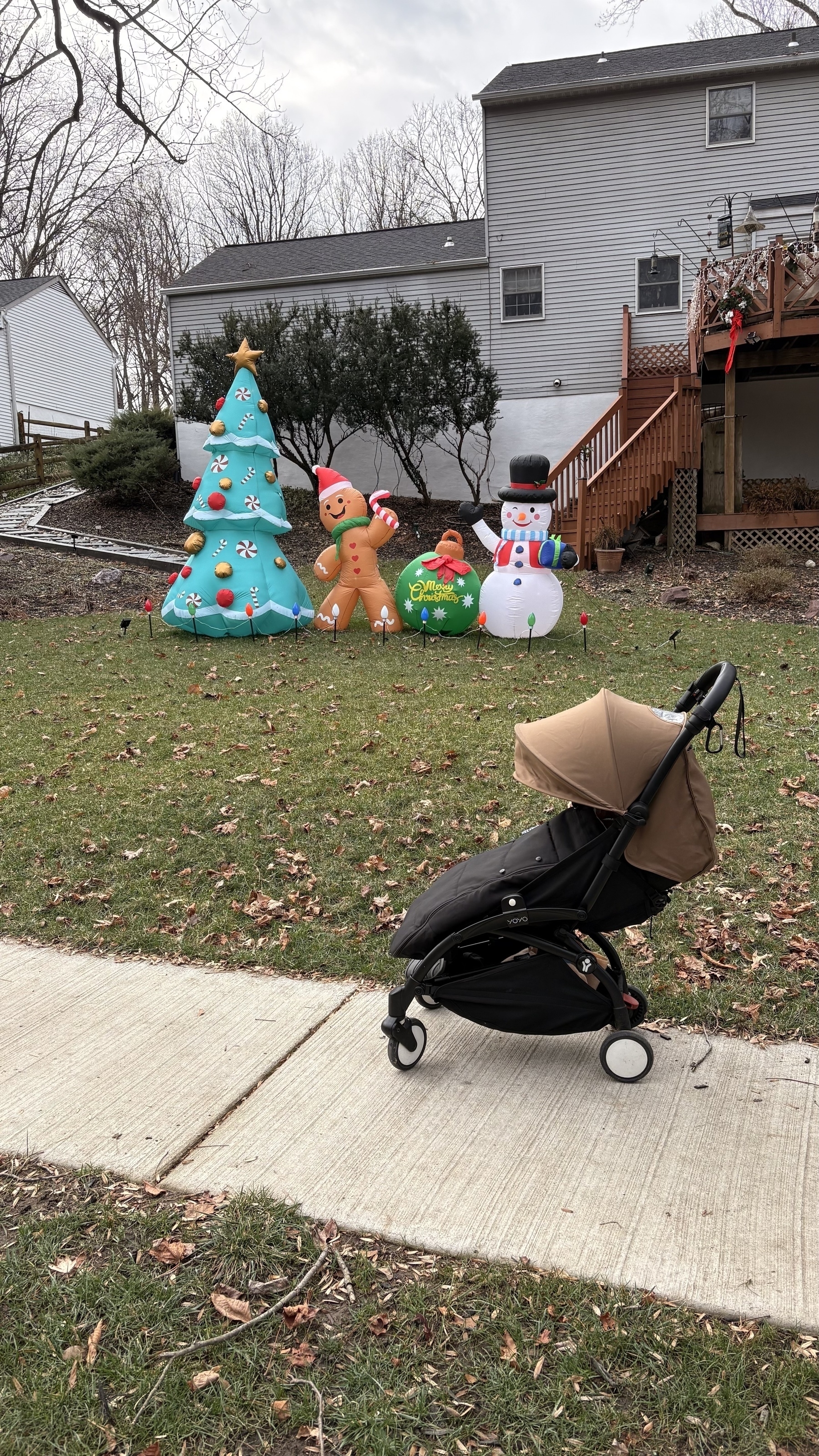 A stroller is parked on a sidewalk in front of a yard with inflatable Christmas decorations, including a tree, gingerbread figures, and a snowman.