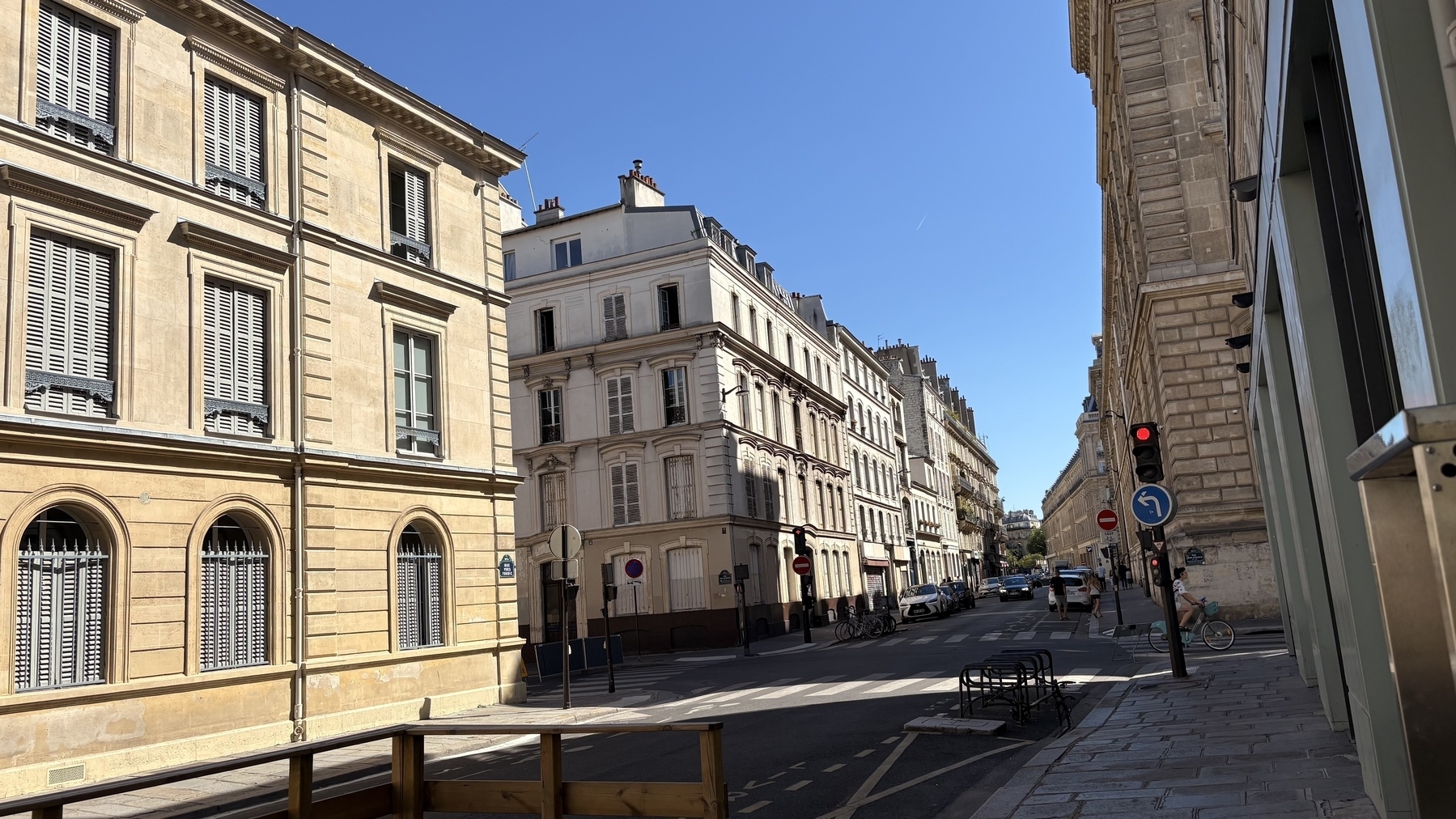 A quiet street lined with classic European-style buildings basks in the bright sunlight under a clear blue sky.