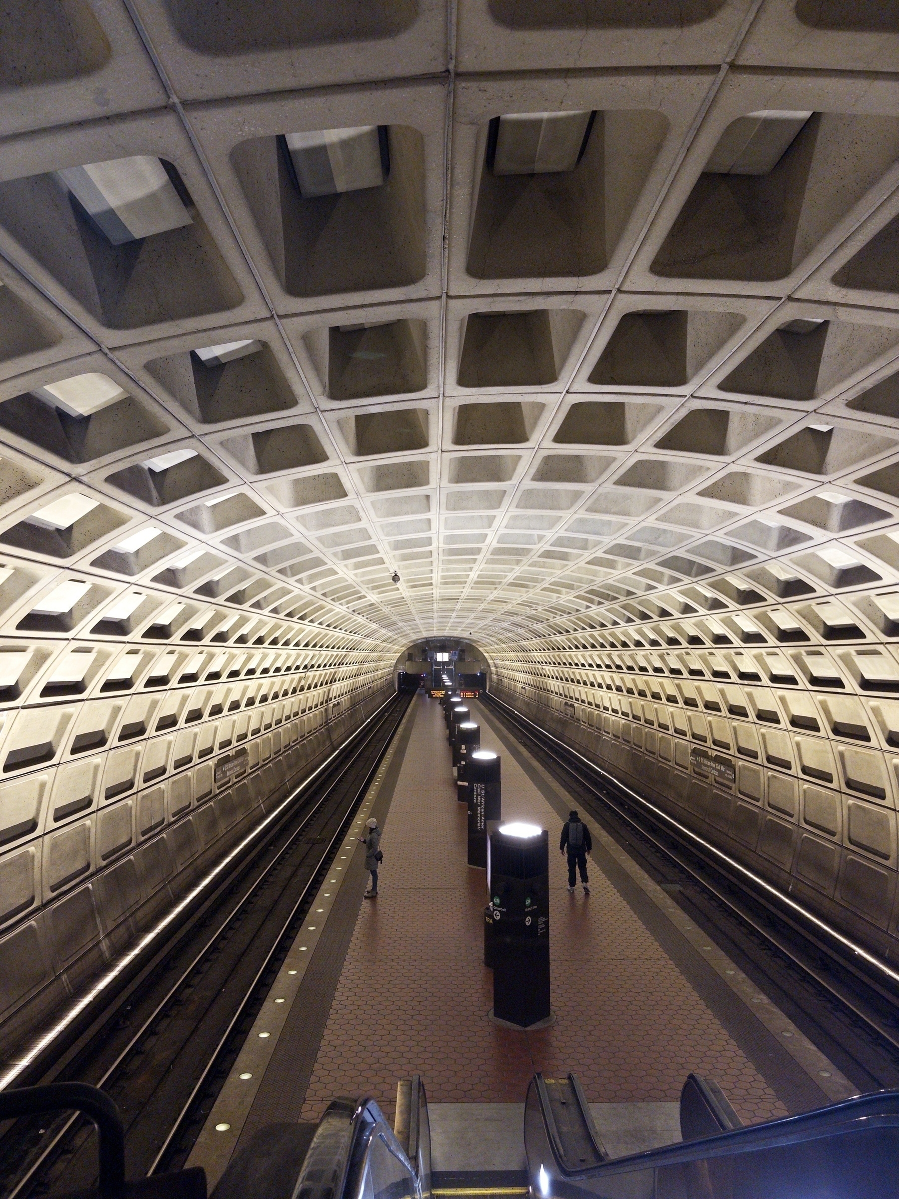 A wide, futuristic-looking metro station features an arched ceiling with geometric patterns, escalators, and tracks flanked by platforms with a few people walking.