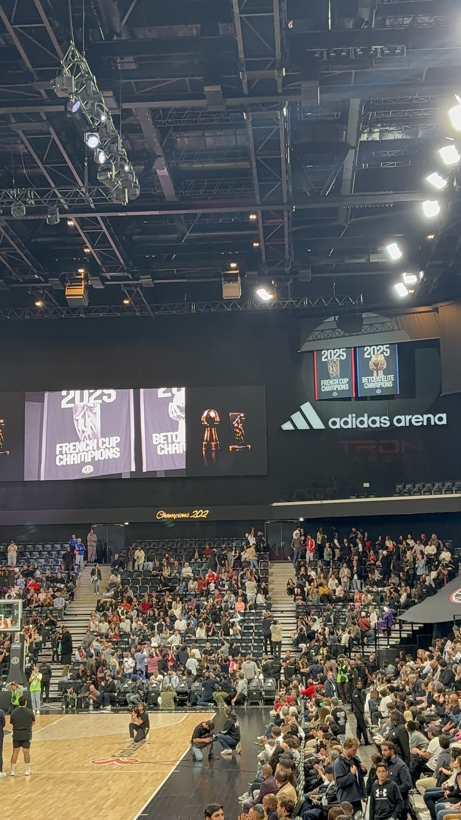 A sports arena is filled with a large crowd, featuring banners and Adidas branding prominently displayed.