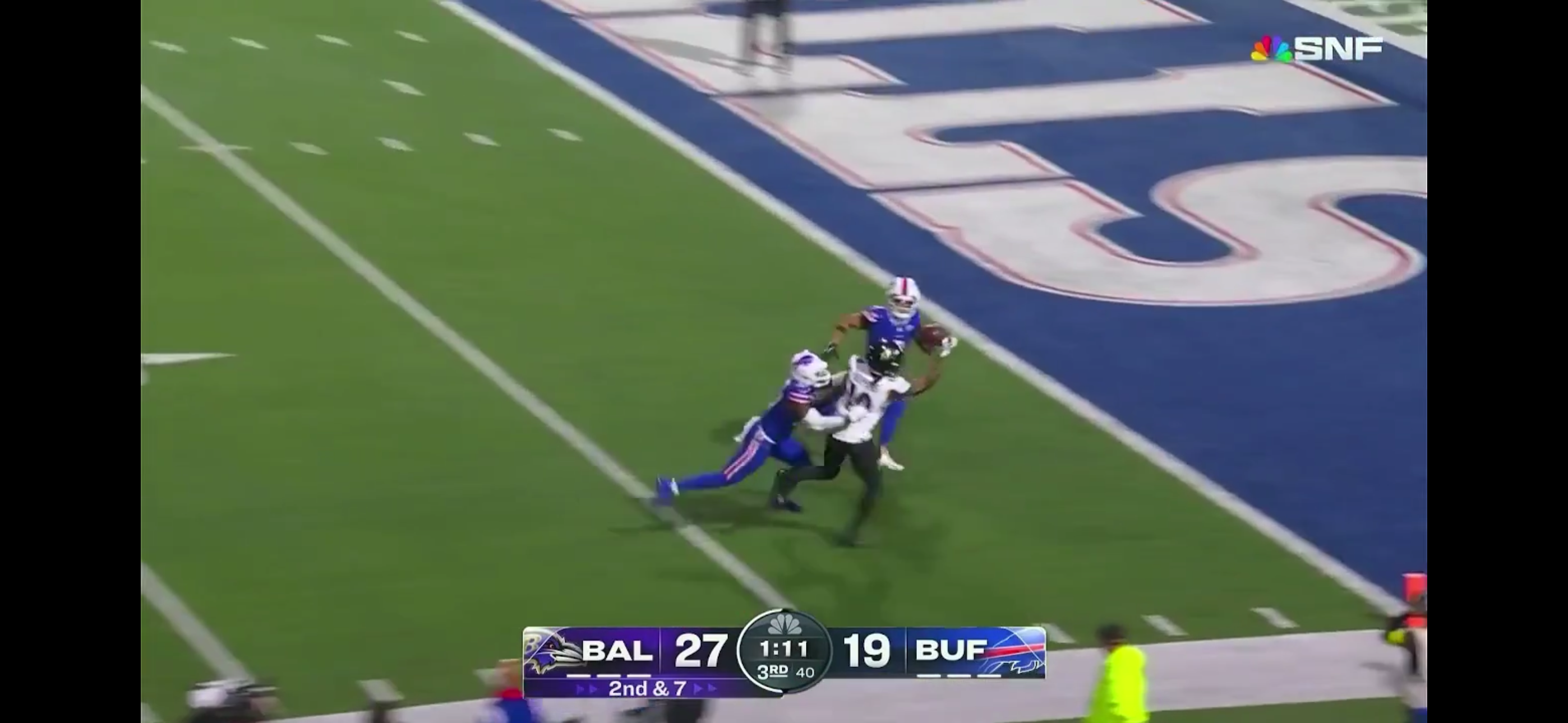 A football player from the Baltimore team attempts to evade tackles from two Buffalo players near the end zone during a game shown on SNF, with a score of 27-19 in favor of Baltimore.
