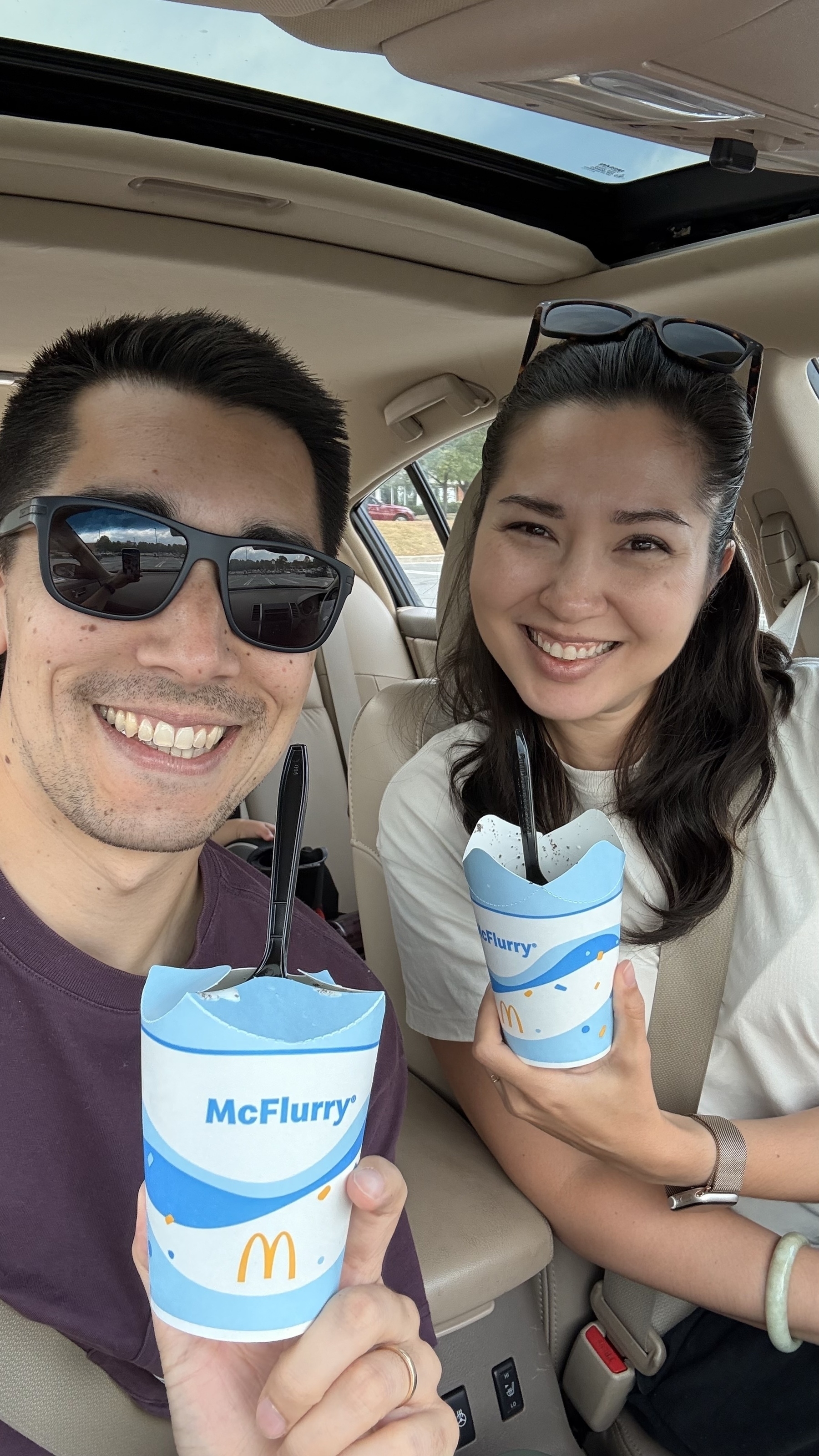 Two people are sitting in a car, smiling, and holding McFlurry desserts.