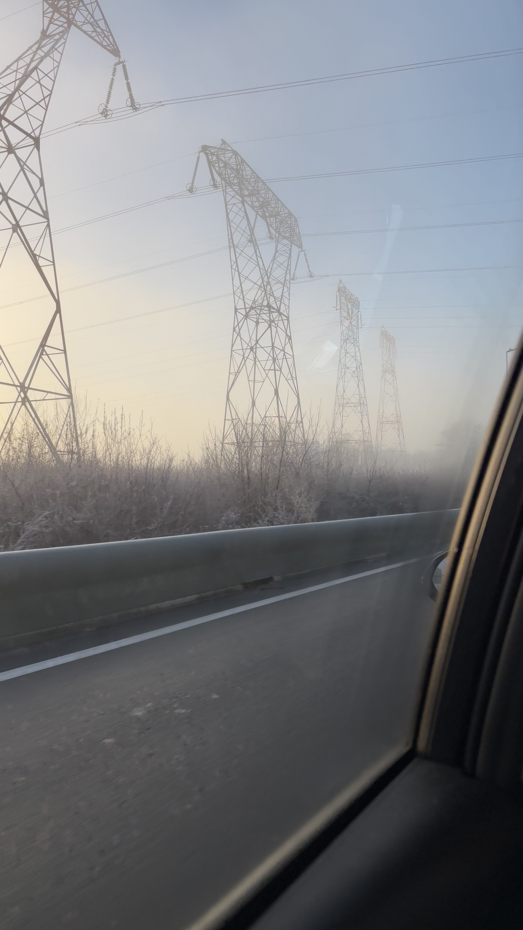 A road runs parallel to a row of power lines and tall electricity pylons surrounded by frosty vegetation under a misty sky.