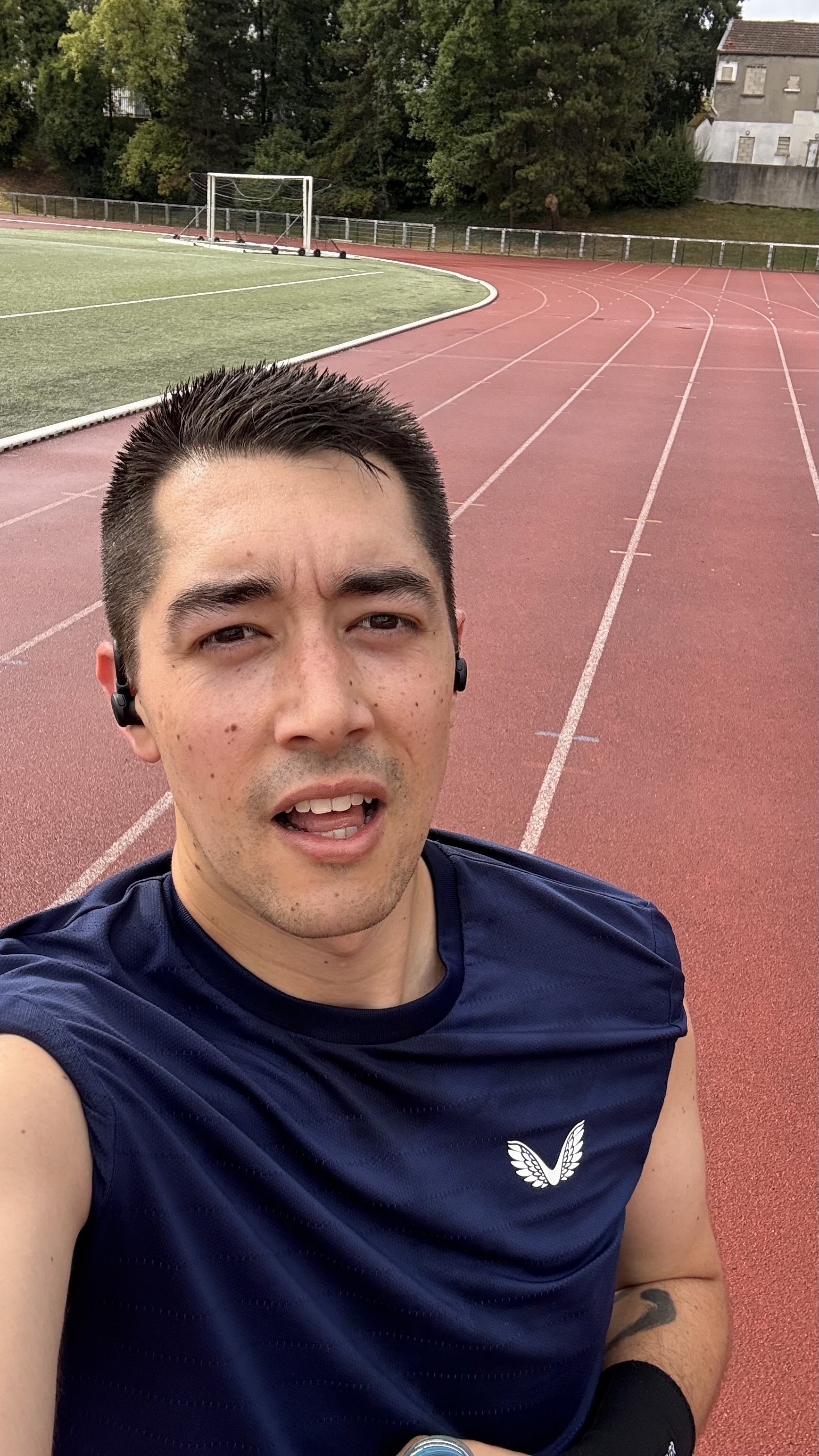 A person wearing a navy blue sleeveless shirt and earbuds is jogging on a red outdoor running track.