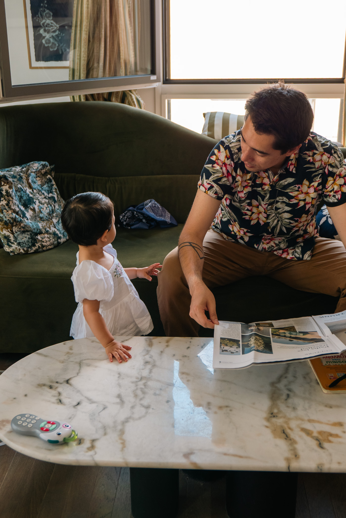 A man wearing a floral shirt is seated on a couch, interacting with a young child standing beside a marble table with items on it.