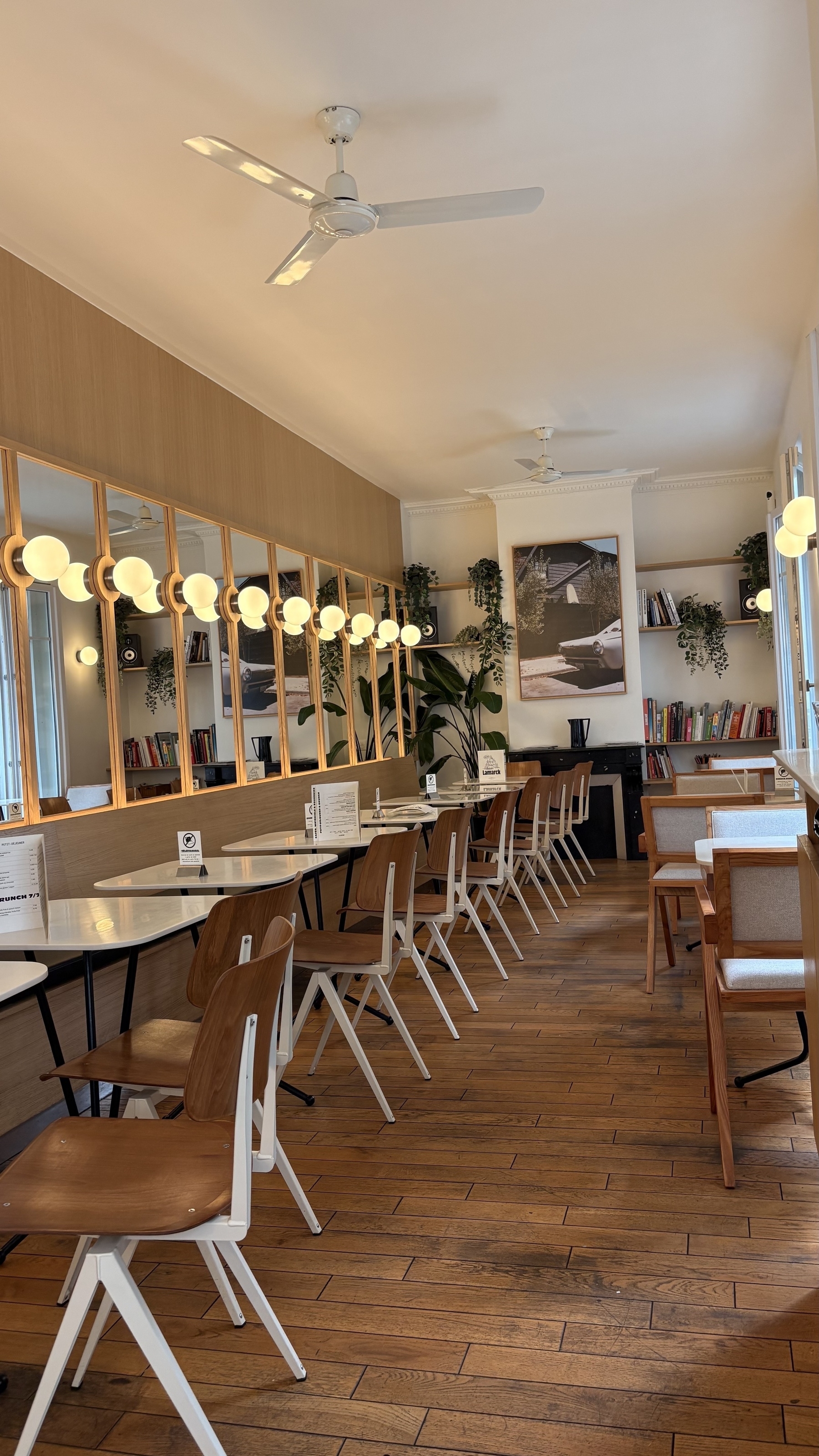 A cozy café features wooden flooring, modern chairs, round tables, and warm globe lights, with plants and framed art for decoration.