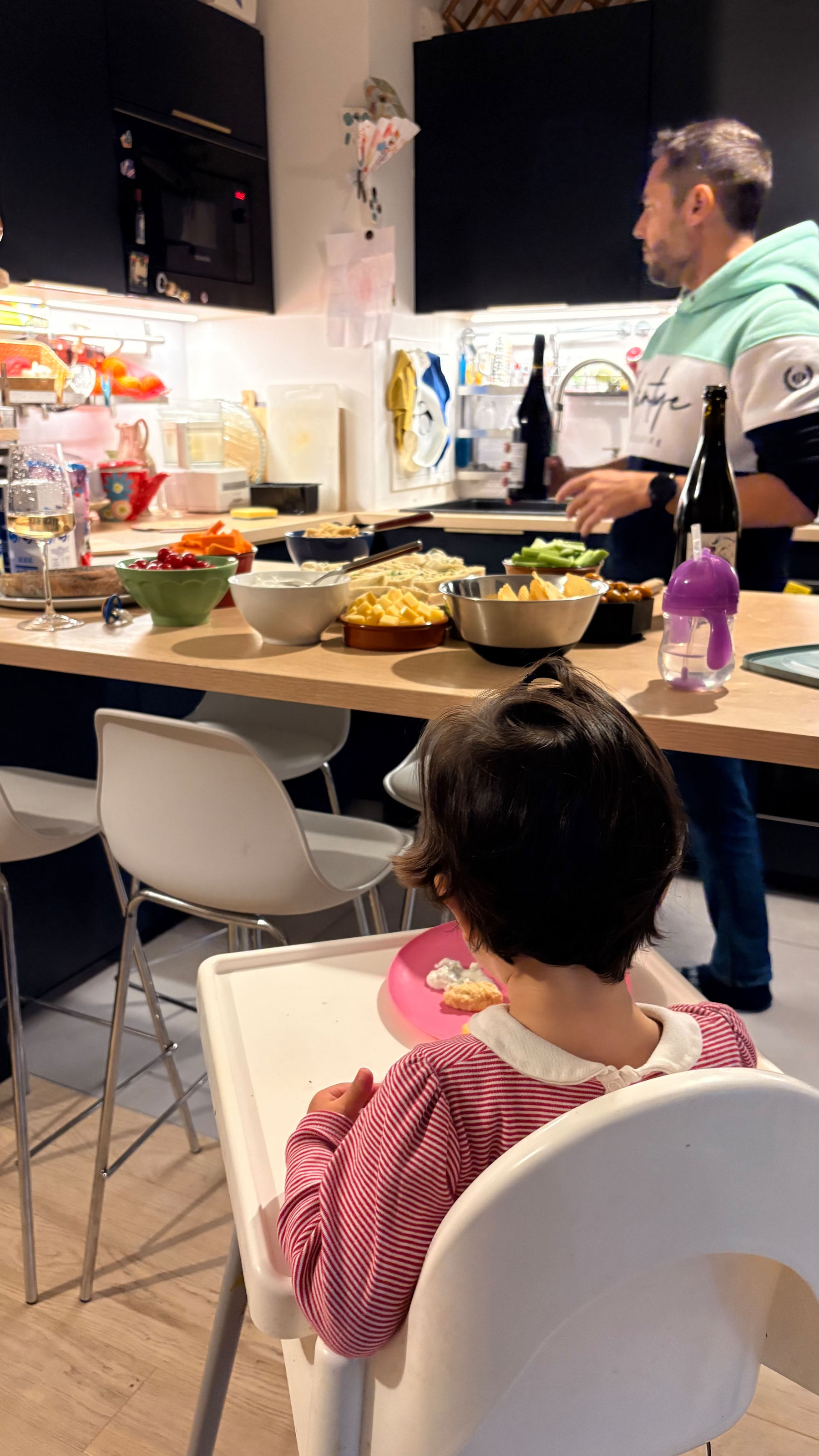 A toddler sits in a high chair eating while a man prepares food at a kitchen counter filled with dishes and bottles.
