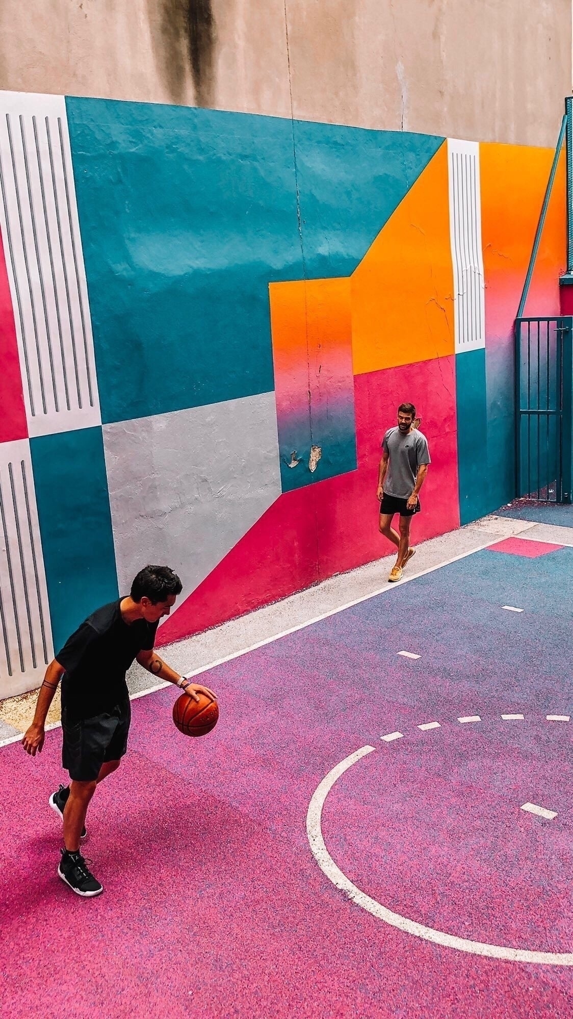 Two people are playing basketball on a vibrantly colored outdoor court with a geometric mural.