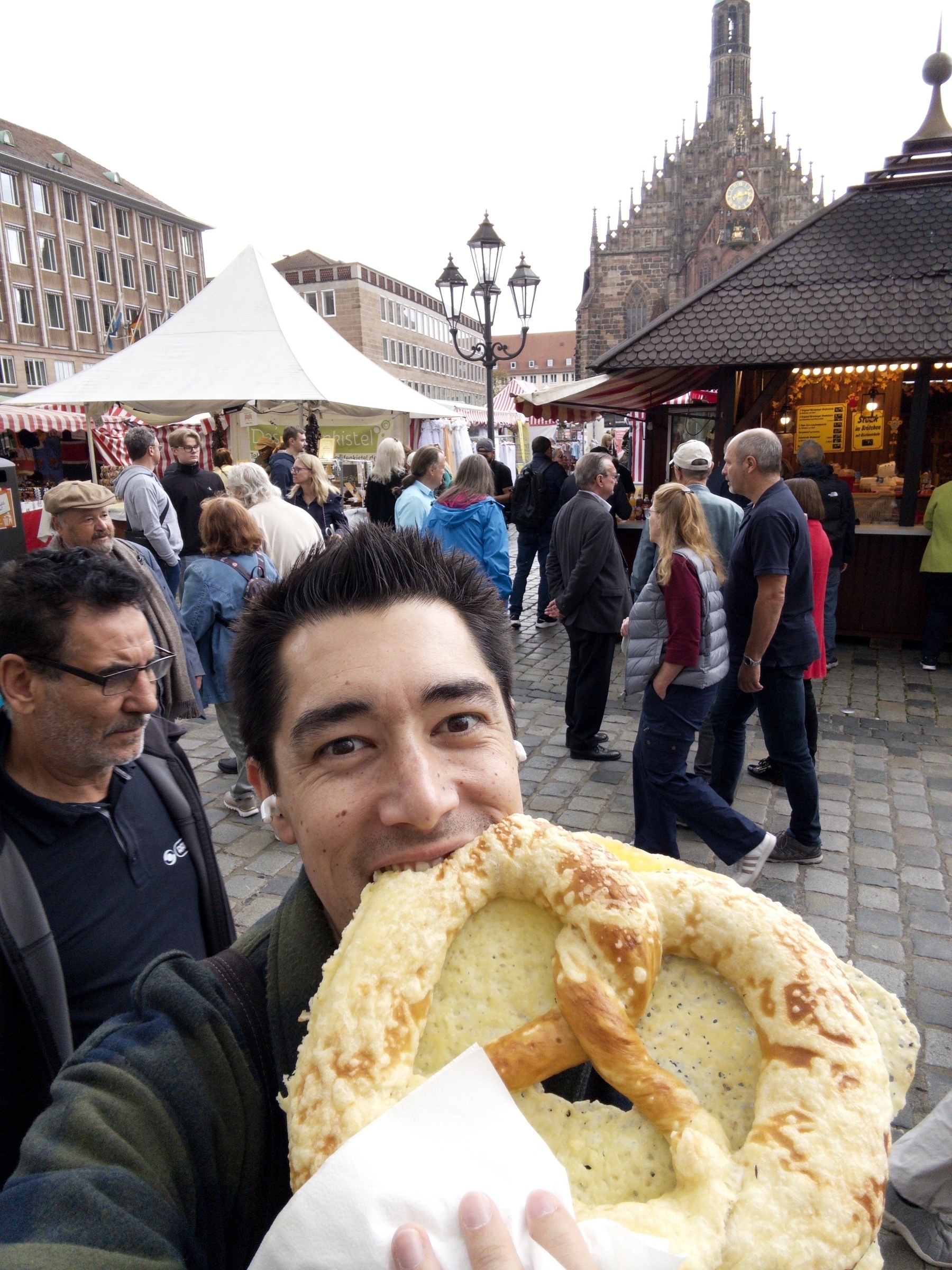 A person is enjoying a large pretzel in a bustling outdoor market with a historical building in the background.