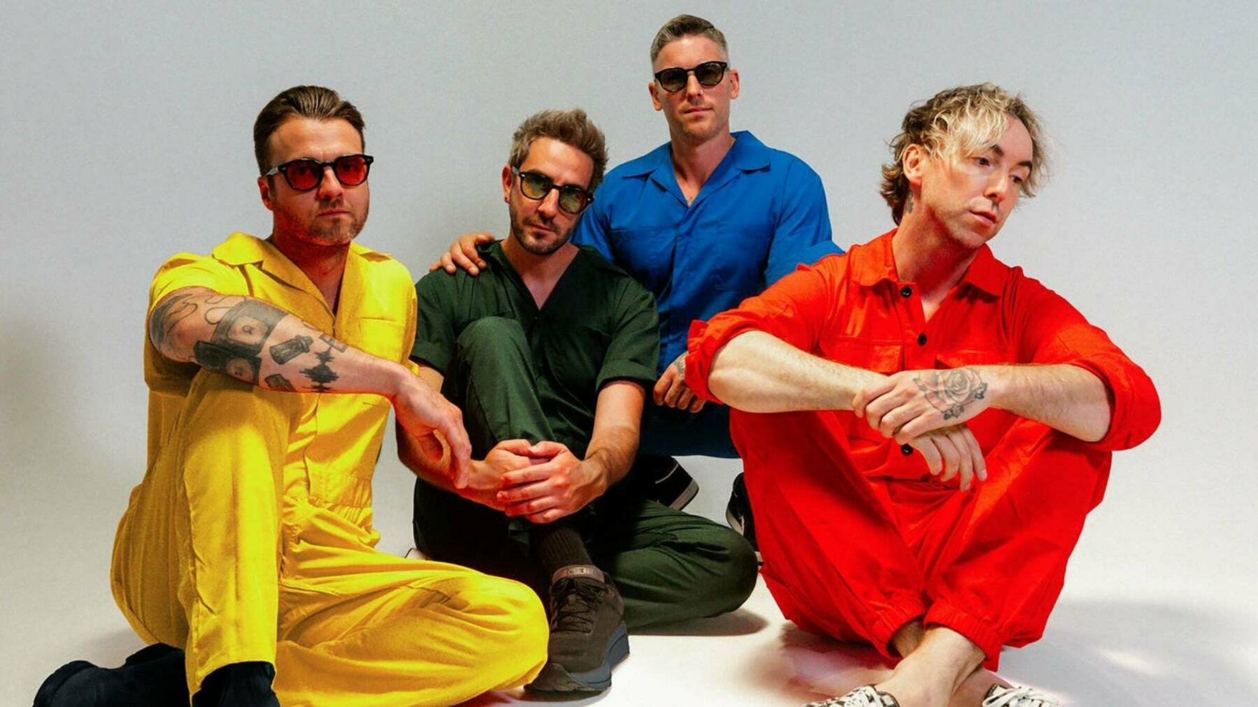 the 4 members of the band All Time Low, wearing colorful jumpsuits and sunglasses are sitting and crouching on the floor against a plain background.