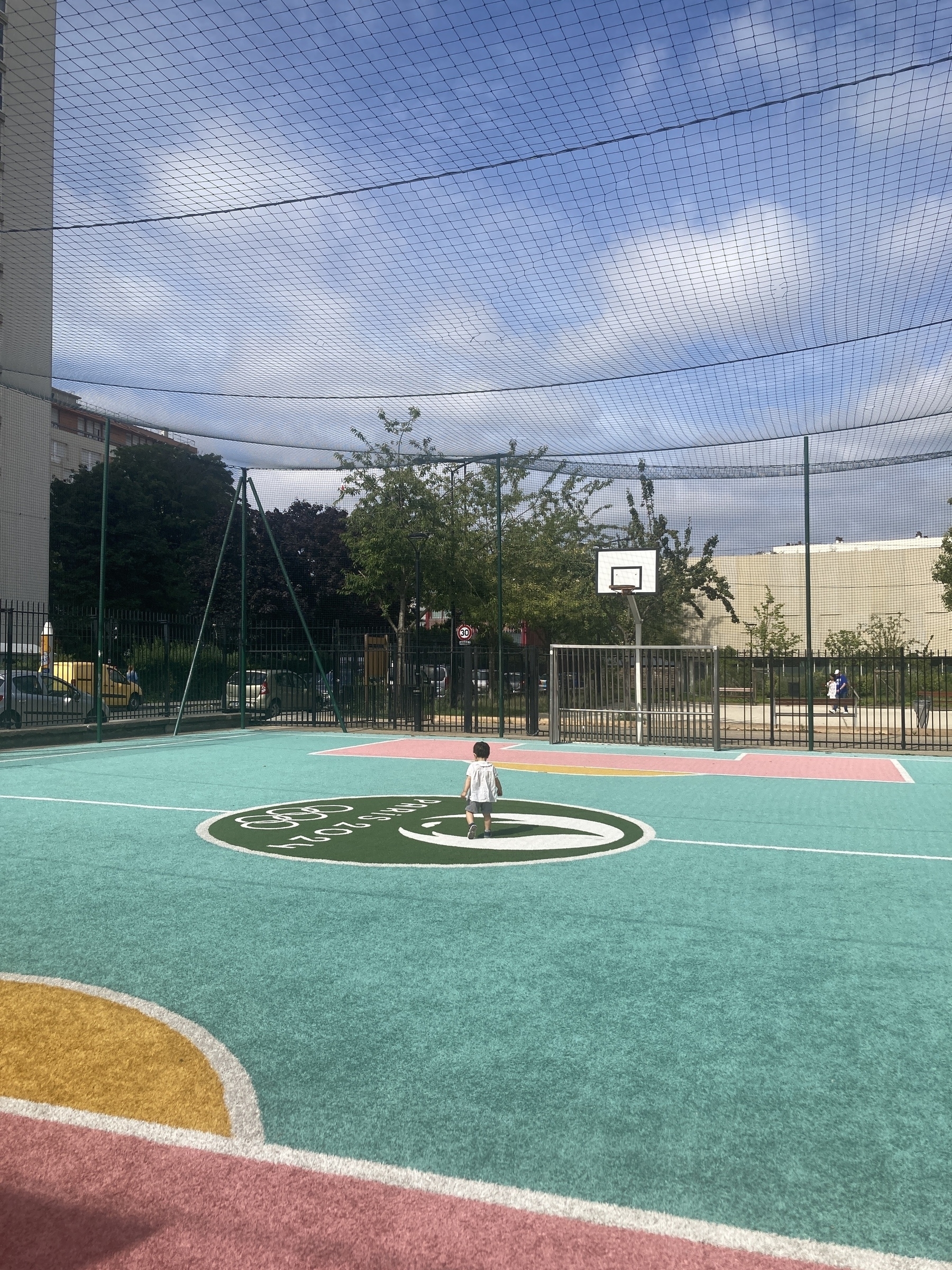 A child stands alone on a colorful, outdoor sports court under a clear sky, surrounded by a fence and trees.