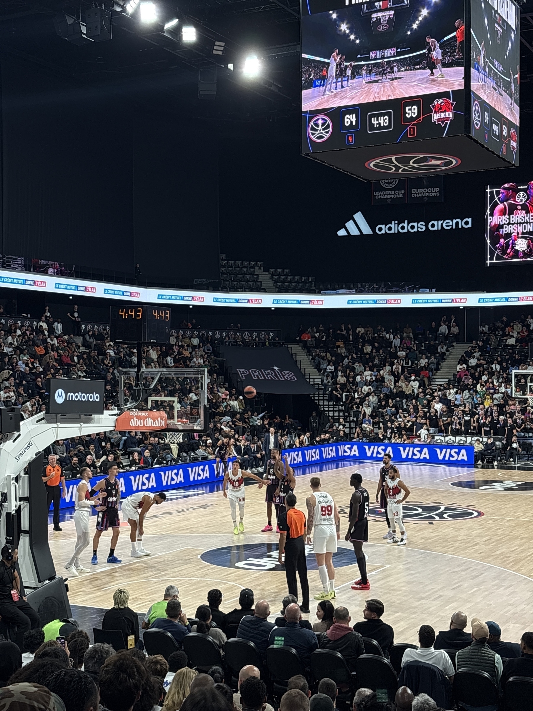 A basketball game is taking place in a packed arena with players on the court and a scoreboard displayed above.