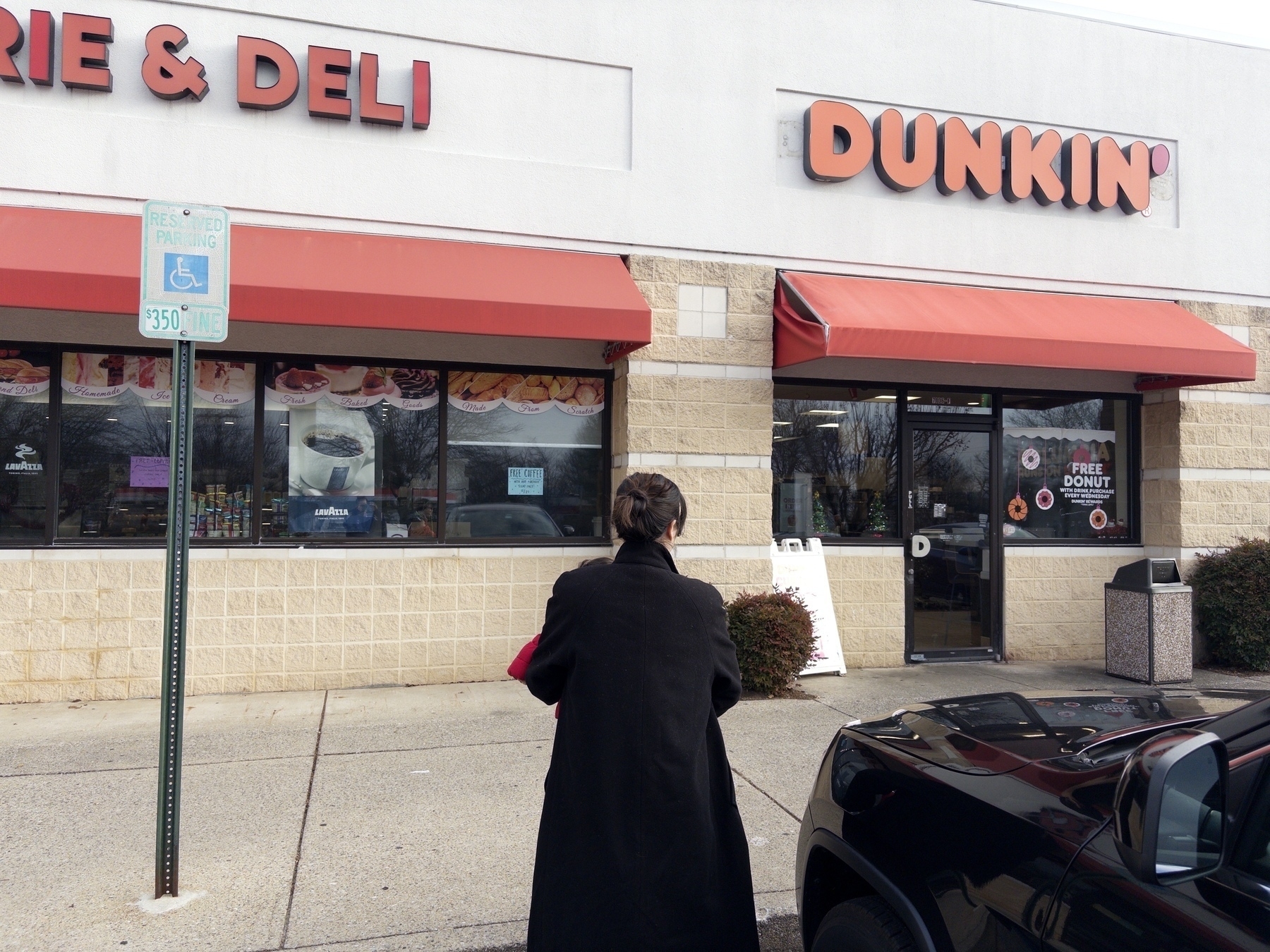 A person is standing in front of a Dunkin' store near a parking area.