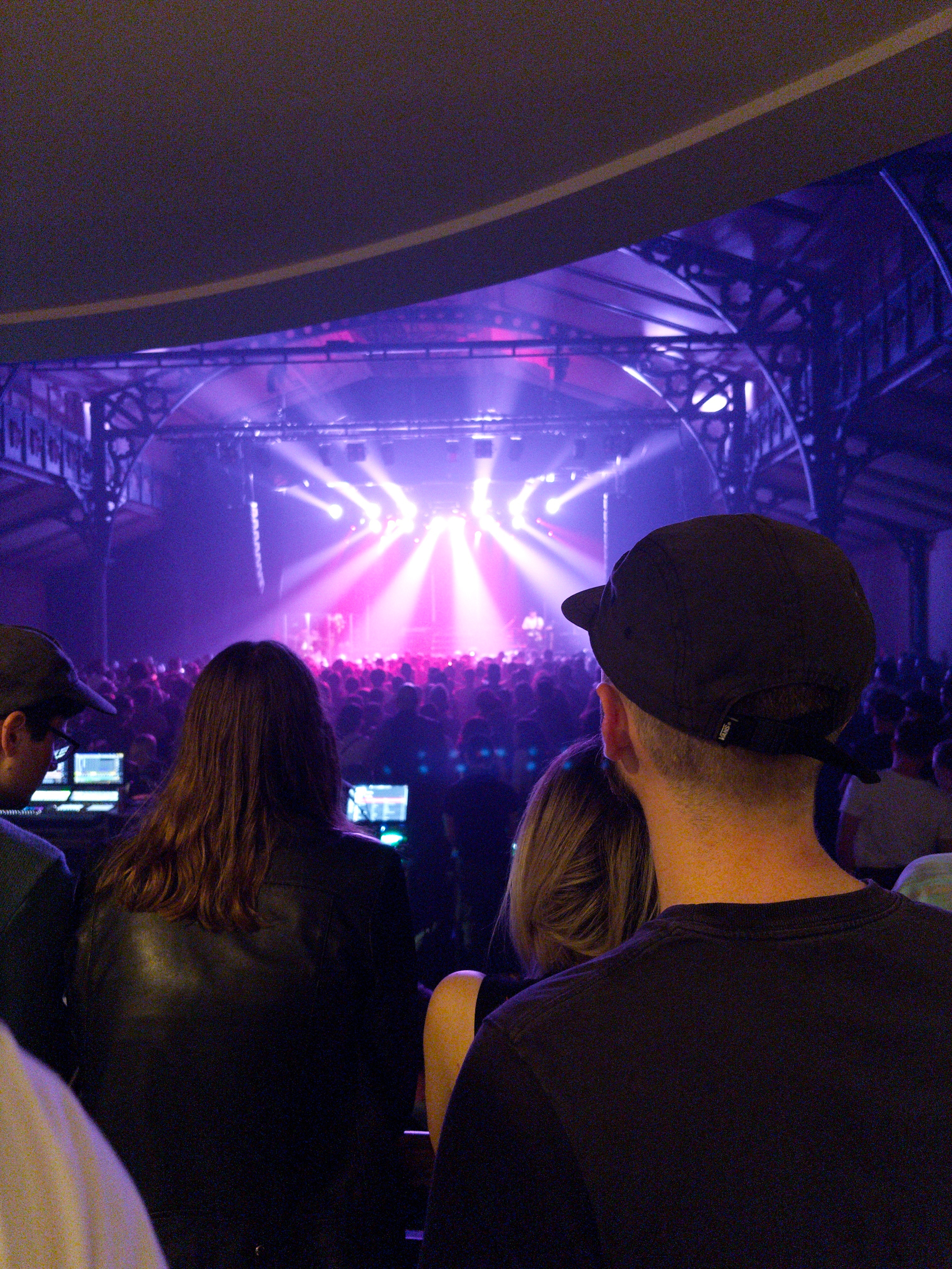 A crowd is watching a live music performance on stage in a dimly lit venue, featuring colorful stage lights.