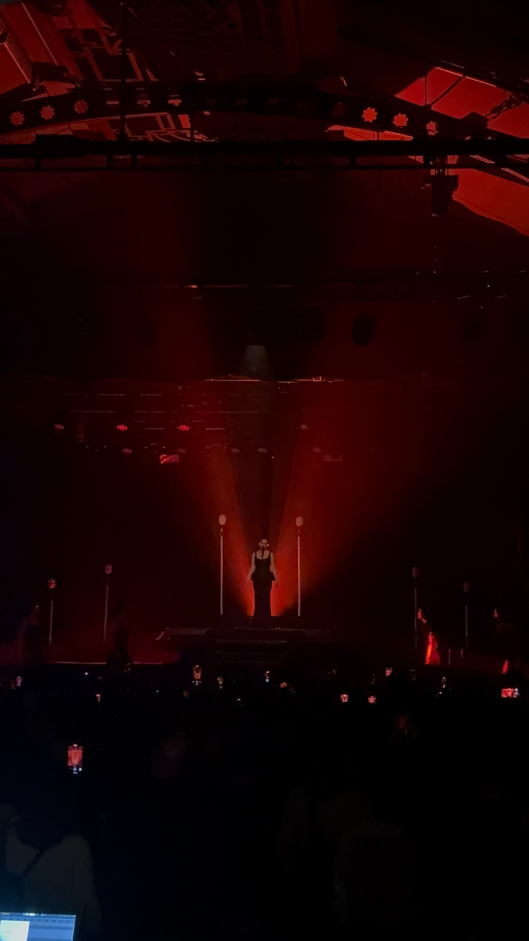 A performer stands on a dimly lit stage under a dramatic red spotlight while an audience watches.