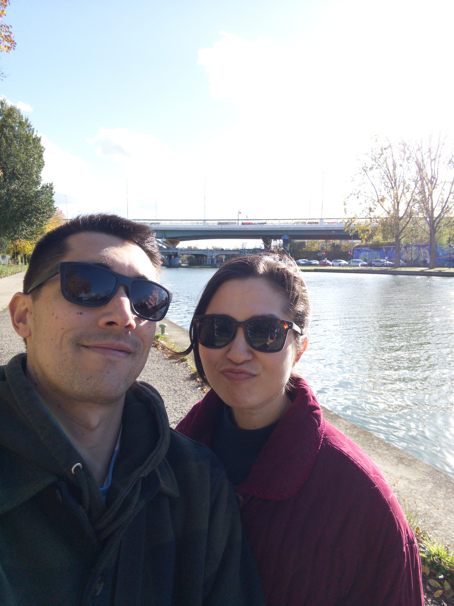 A man and a woman wearing sunglasses are smiling by a river with a bridge in the background.