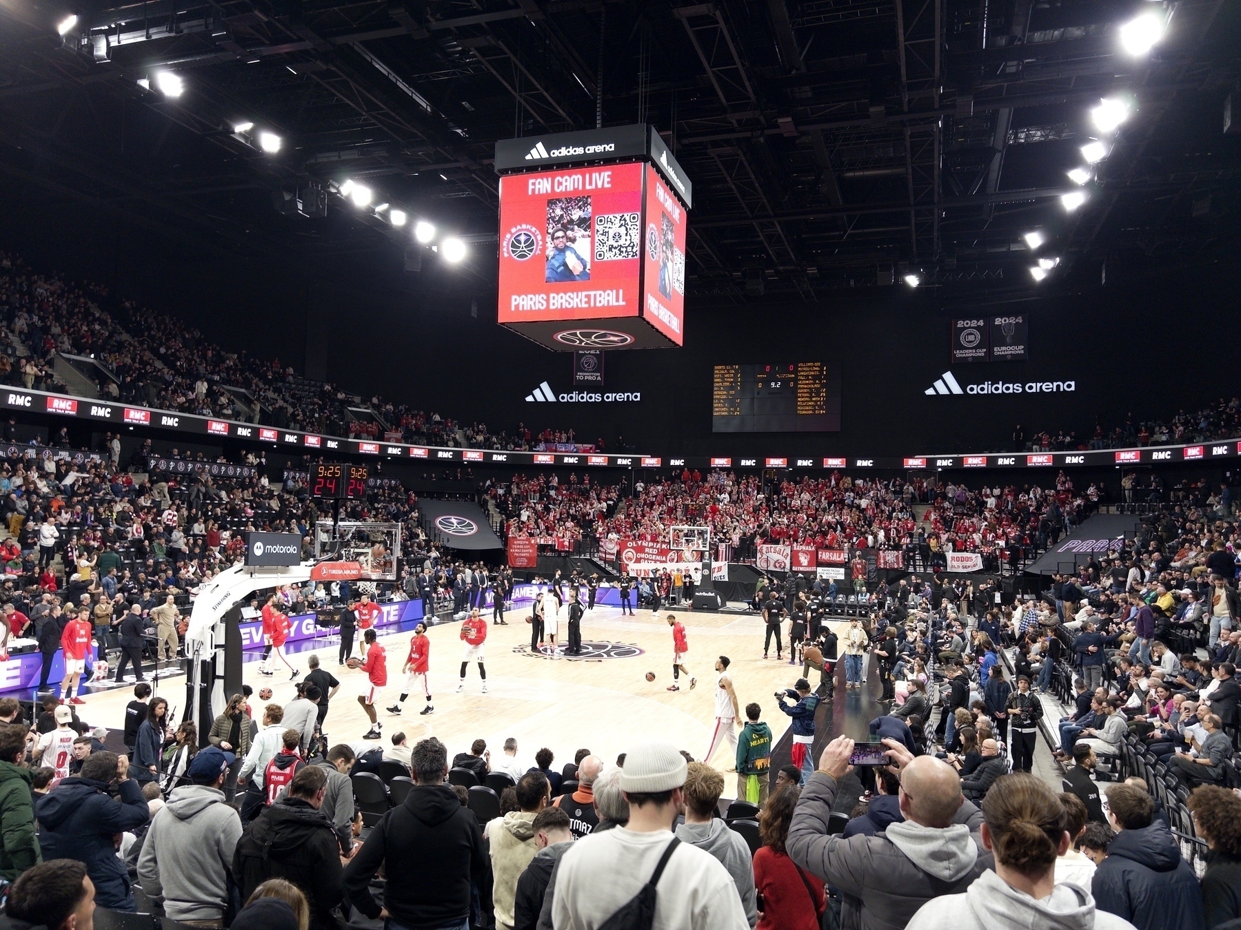 A packed indoor basketball arena features a game in progress with fans filling the stands and banners displayed around the court.