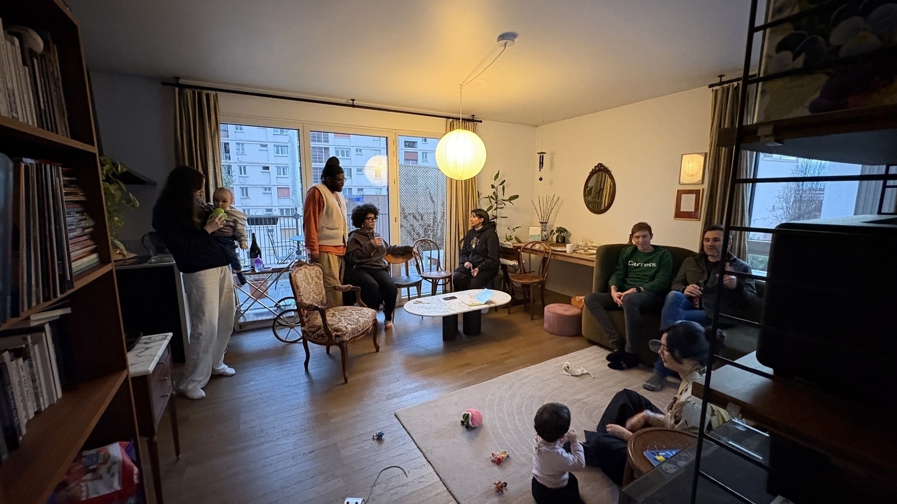 A group of people are gathered in a cozy, well-lit living room, some sitting and some standing, with children playing on the floor.