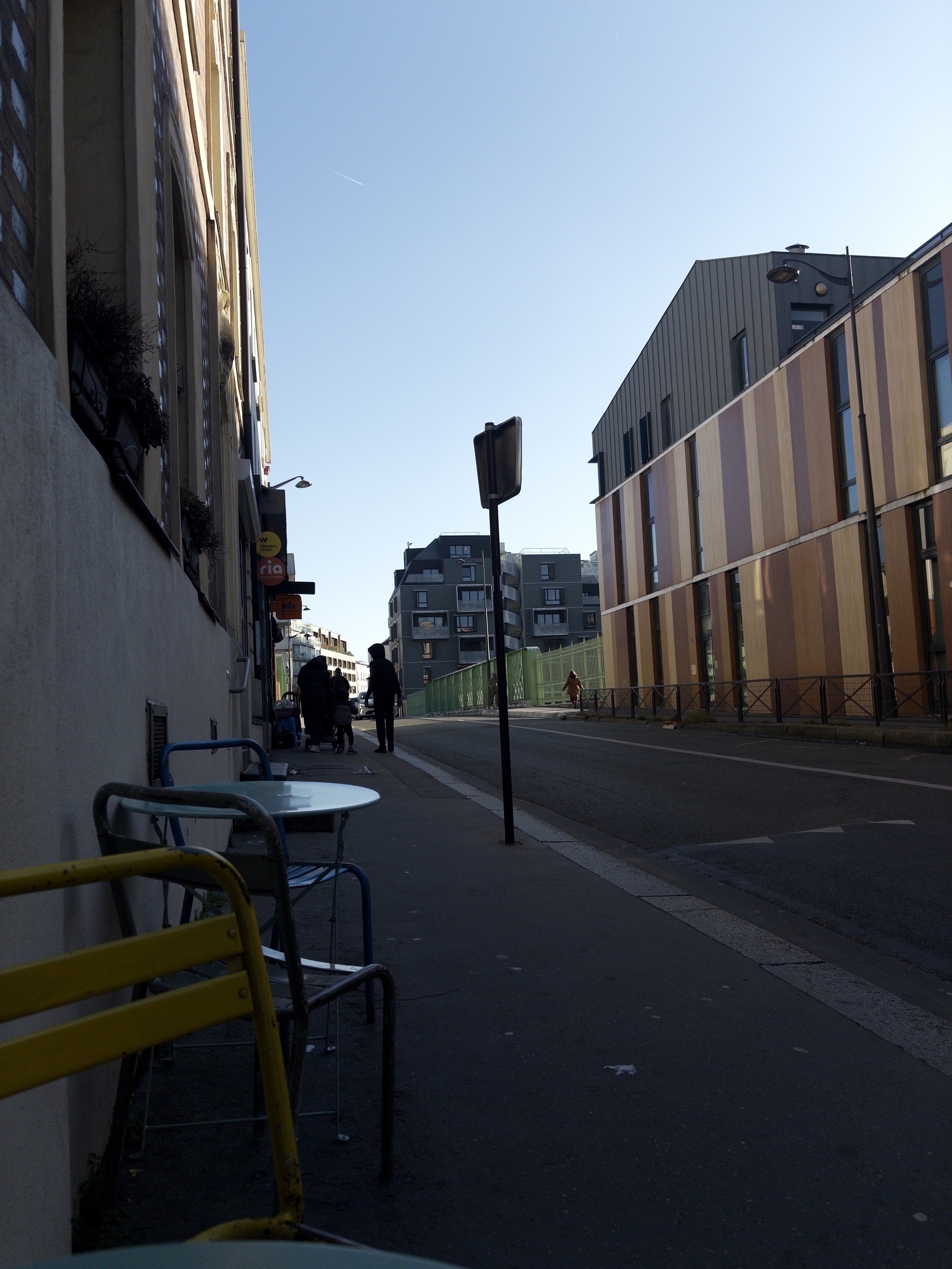 A narrow urban street is lined with modern buildings and a sidewalk featuring a few outdoor tables and chairs.