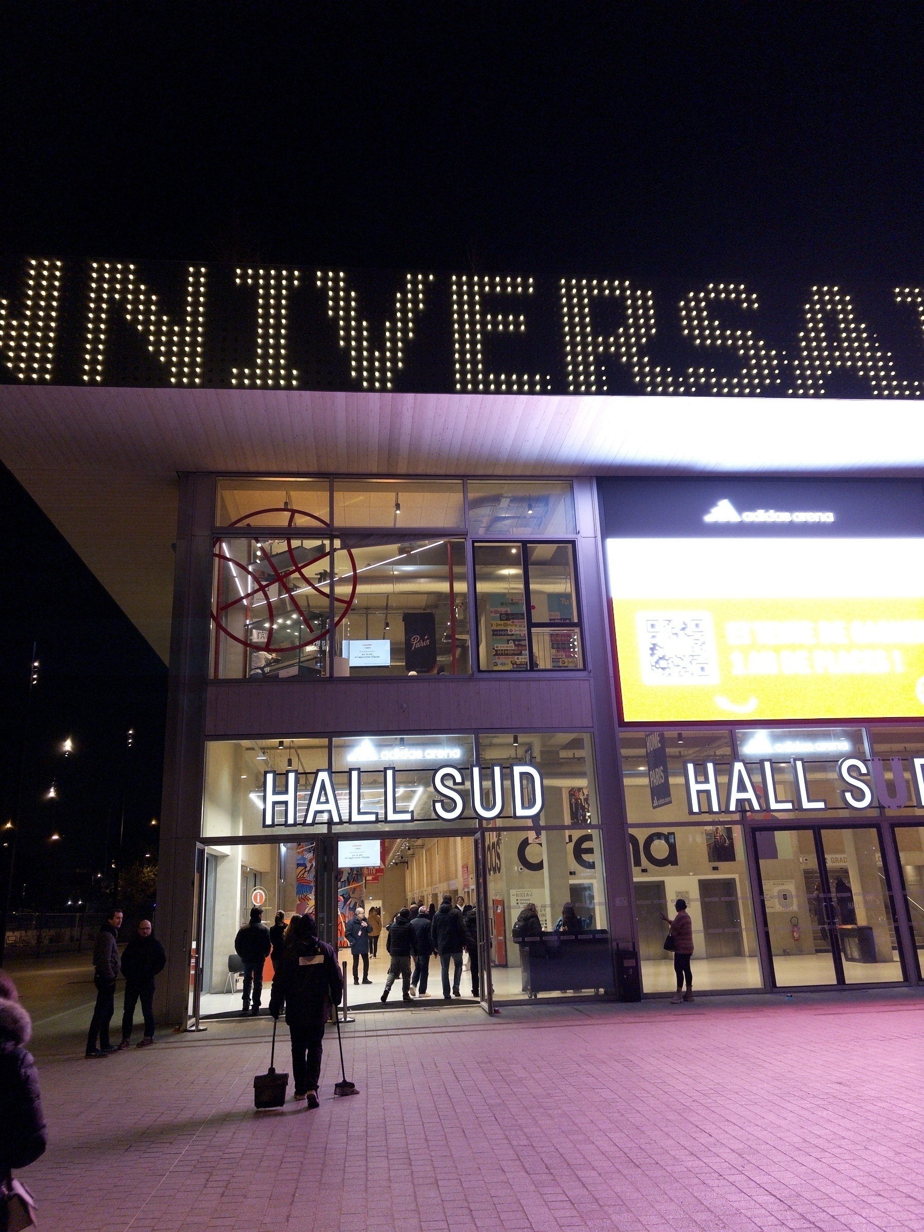 People are gathered outside an illuminated entrance labeled 'HALL SUD,' under a large structure with the word 'ANNIVERSAIRE' displayed in lights.