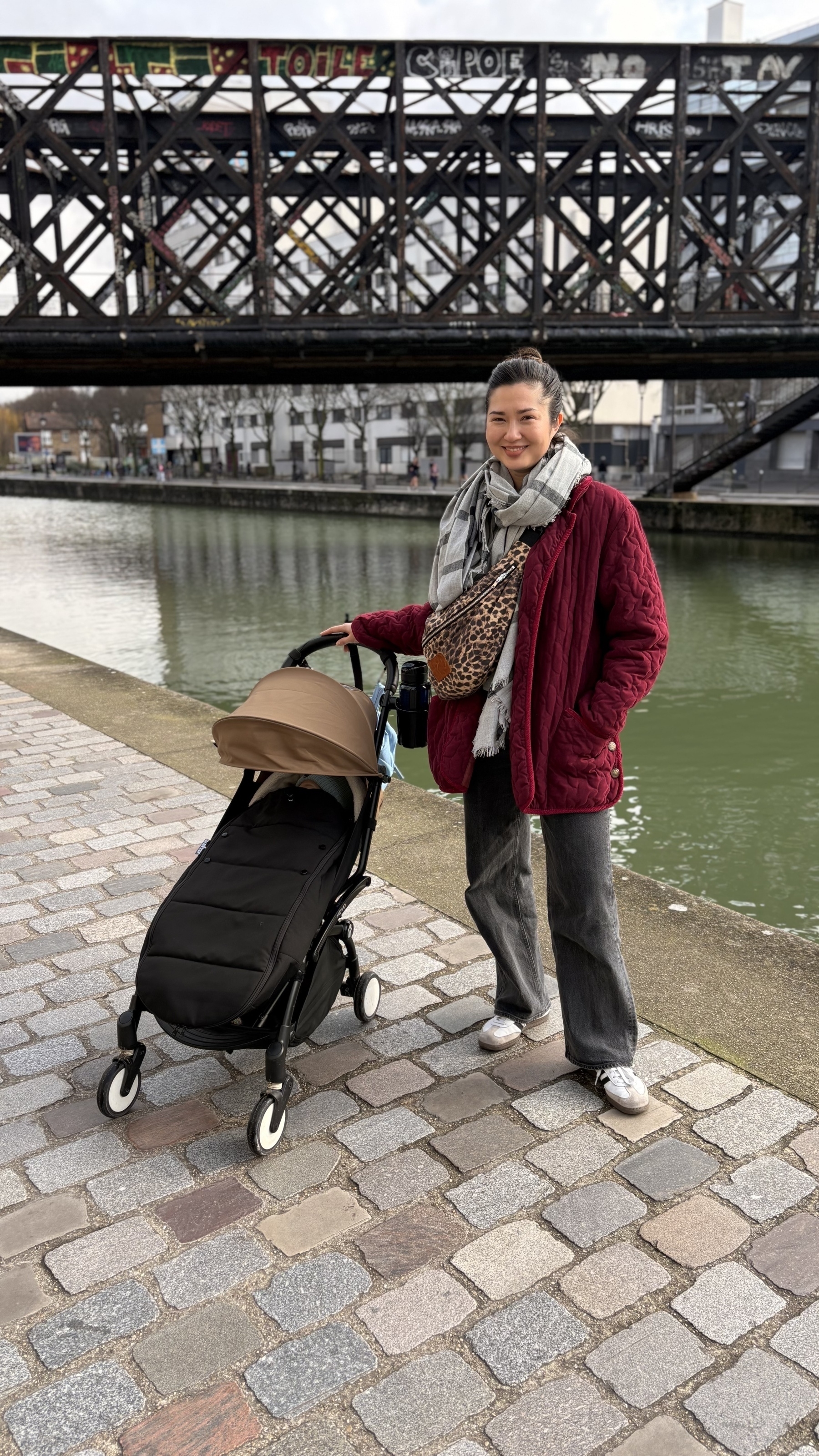 A person in a red coat stands next to a stroller by a canal, with a bridge in the background.