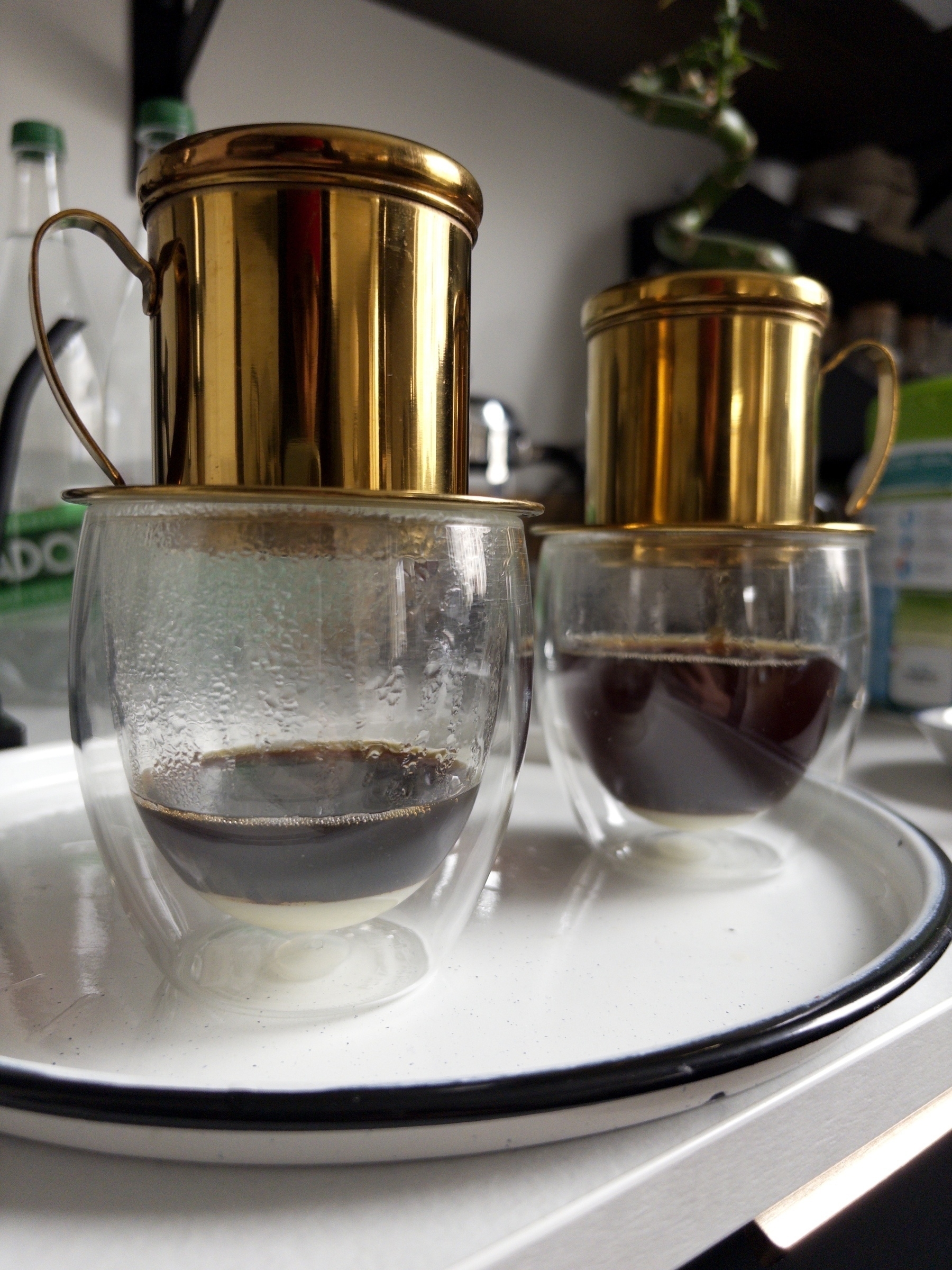 Auto-generated description: Two Vietnamese phin filters drip coffee into double-walled glasses on a white tray.