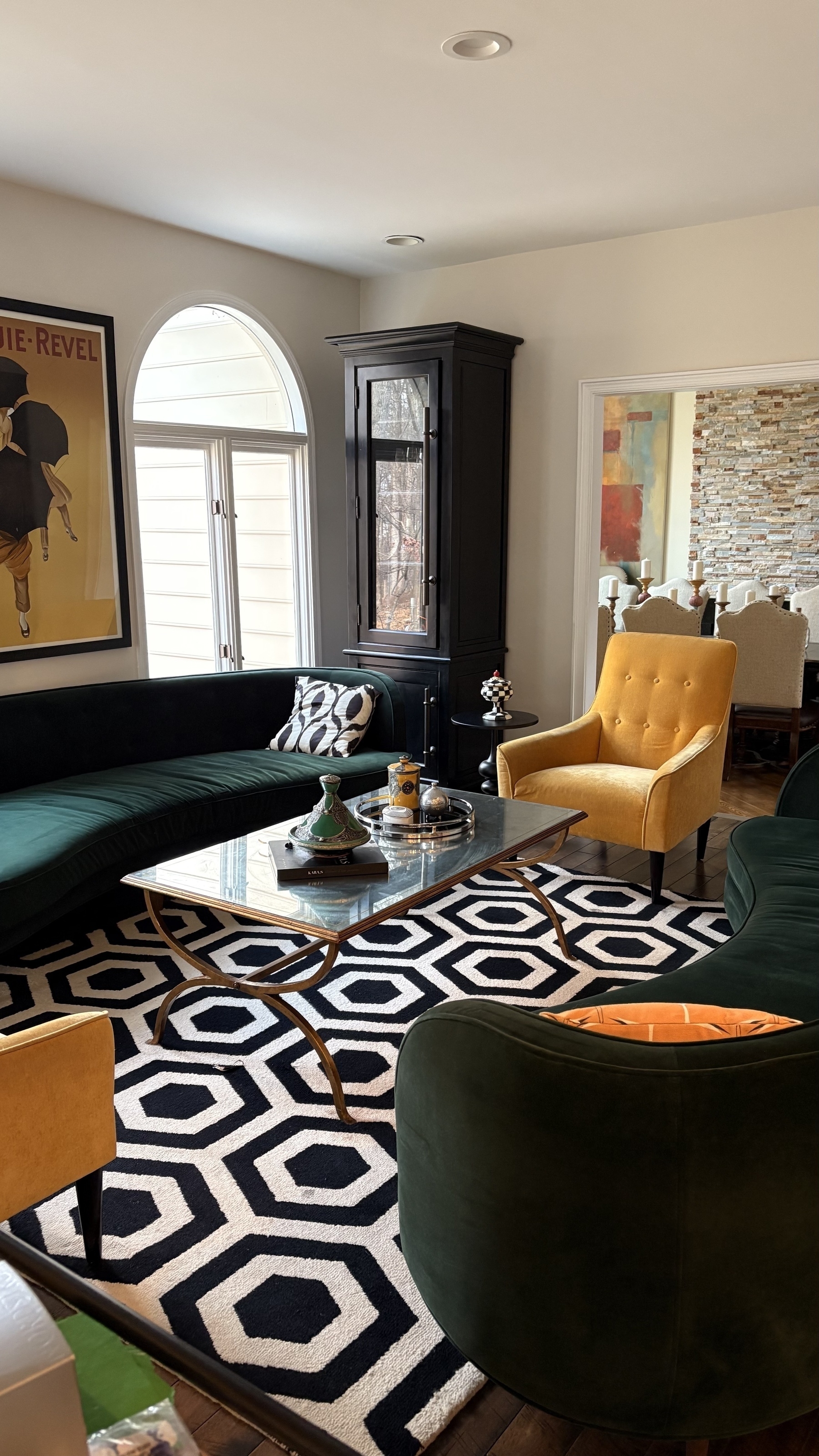 A stylish living room features a green curved sofa, yellow armchairs, a geometric black-and-white rug, and a glass coffee table, with artwork and an elegant dining area visible in the background.