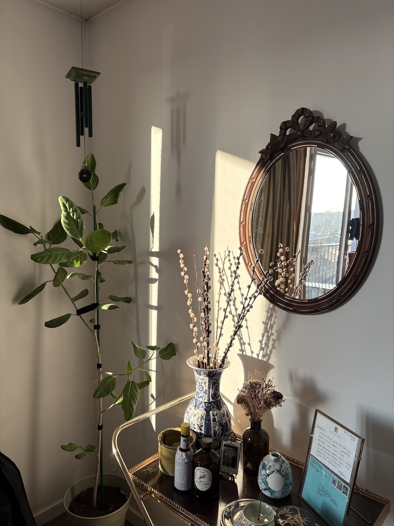 A cozy room corner features a potted plant, a decorative mirror, a vase with dried flowers, and various small decorative items on a table.