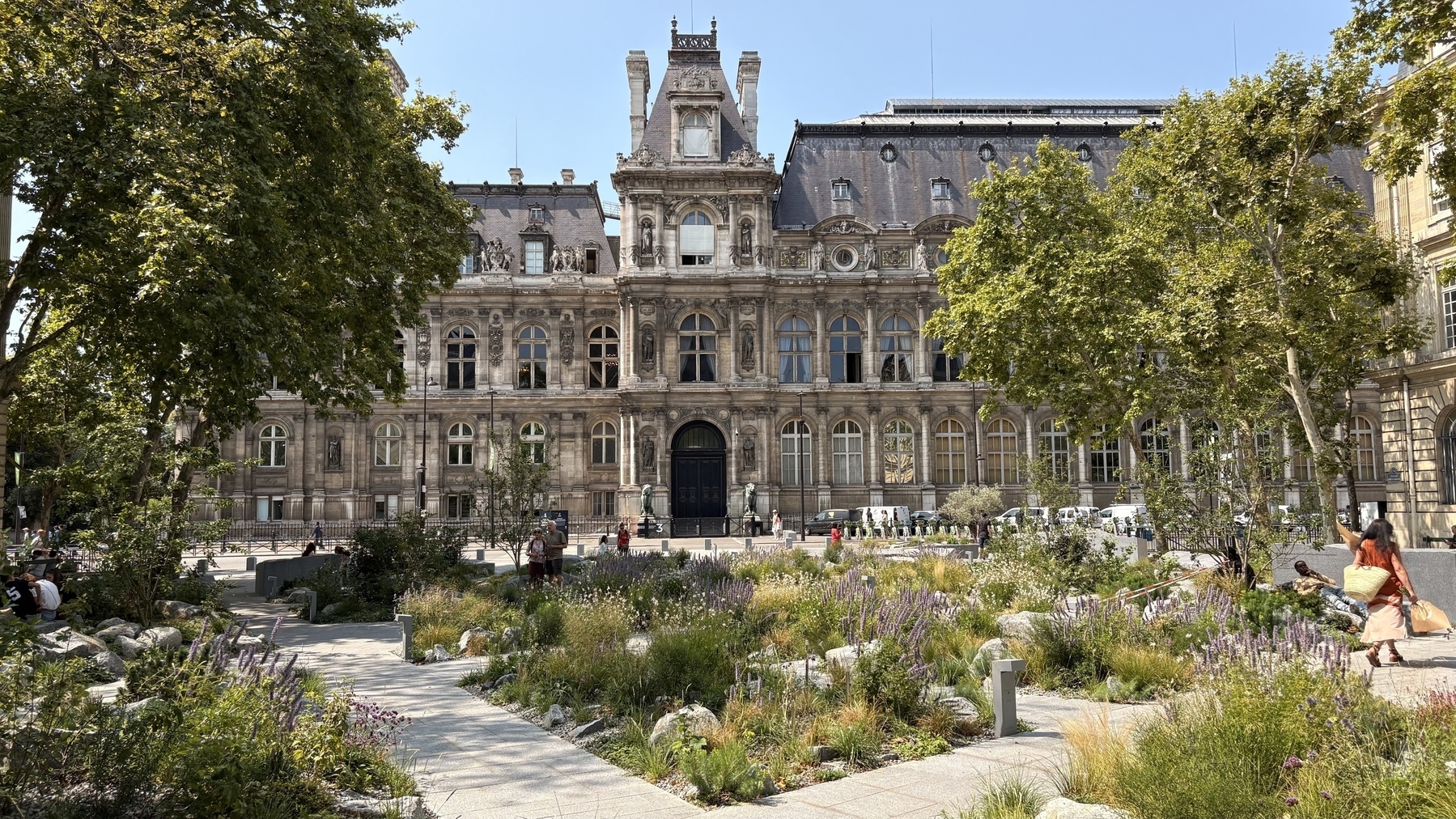 A historic building with ornate architecture is surrounded by a lush garden and trees, with people walking and relaxing.