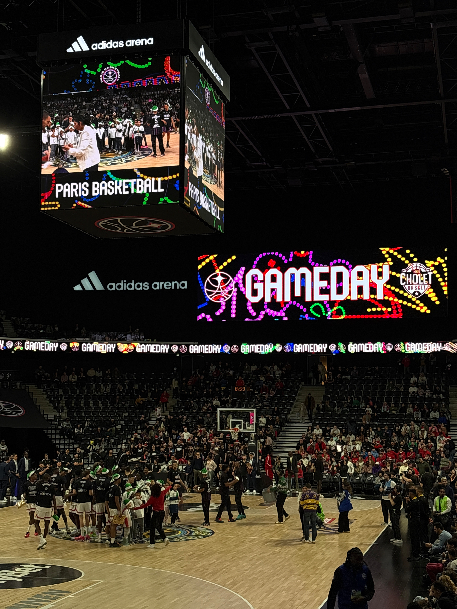A basketball game is taking place in an indoor arena with a Paris Basketball and GAMEDAY display on a large screen.