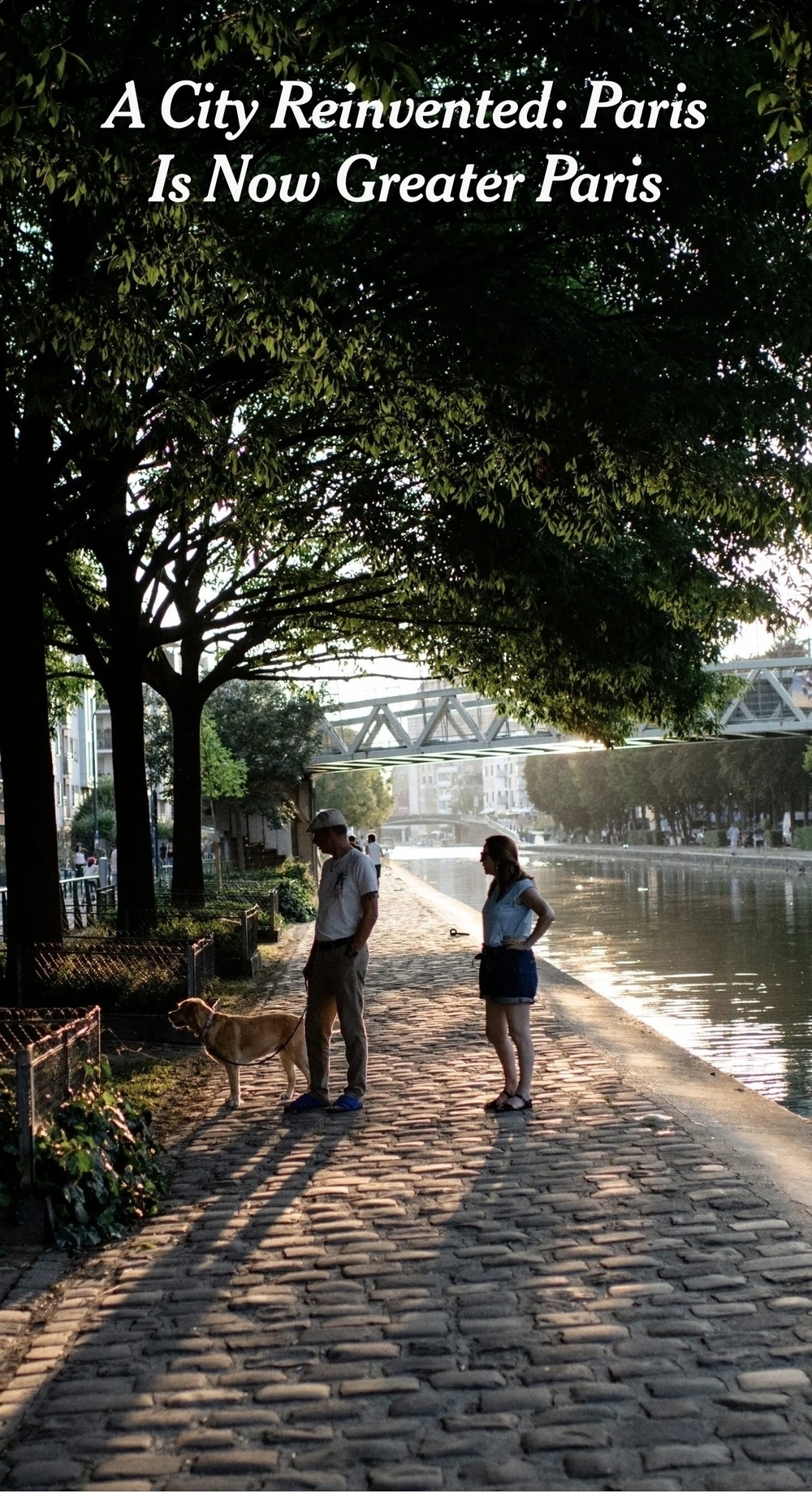 A man and a woman with a dog stand by a cobblestone path next to a river, surrounded by trees, with a bridge in the background.