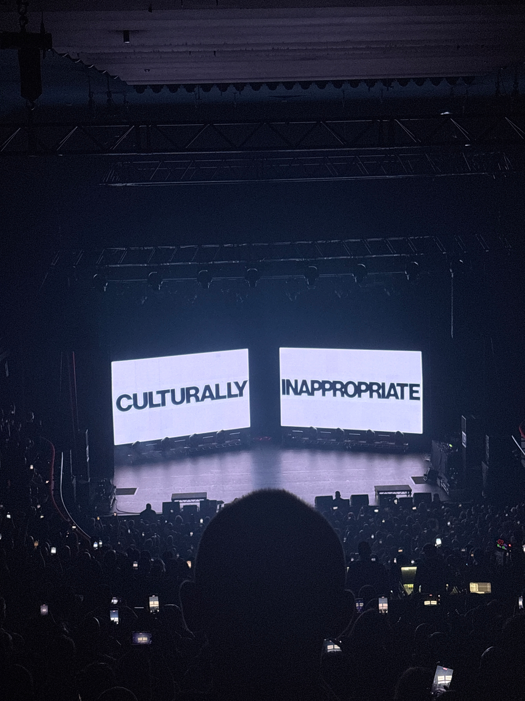An indoor stage is displayed with two large screens showing the words CULTURALLY INAPPROPRIATE to a darkened audience.