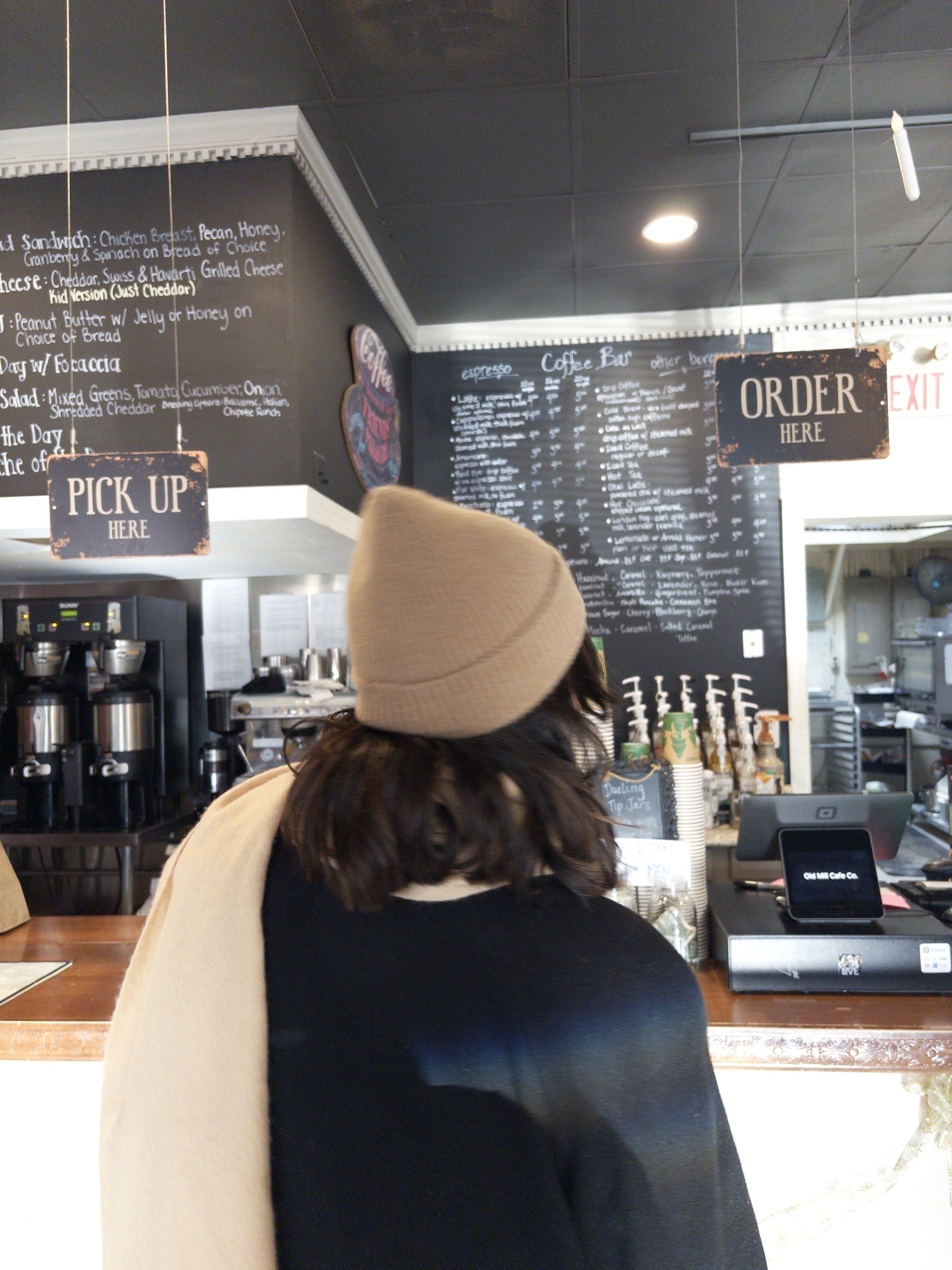 A person wearing a hat stands in front of a café counter with a menu board and coffee equipment.