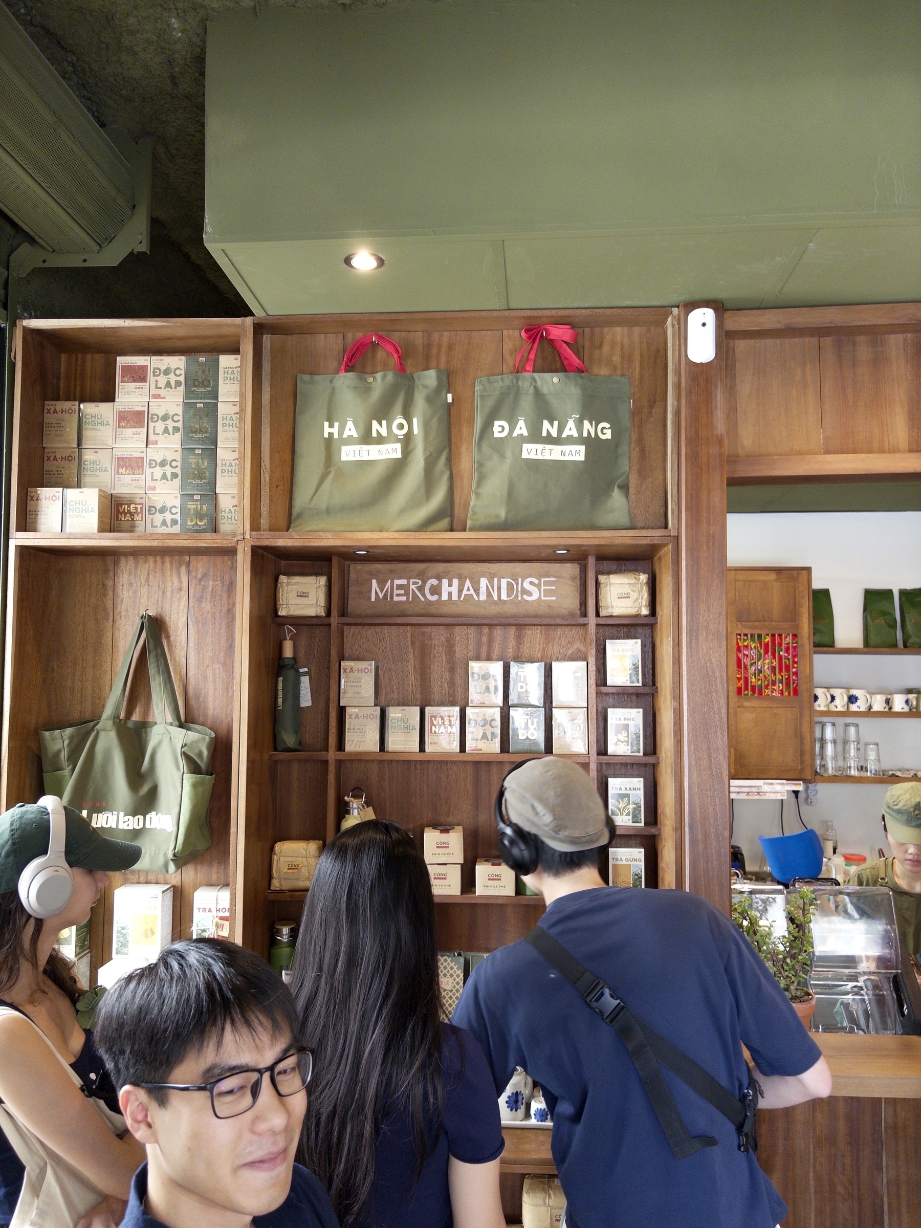 A group of people browse merchandise displayed on wooden shelves, with bags labeled ‘HA NÓI’ and ‘DA NANG’ prominently featured.
