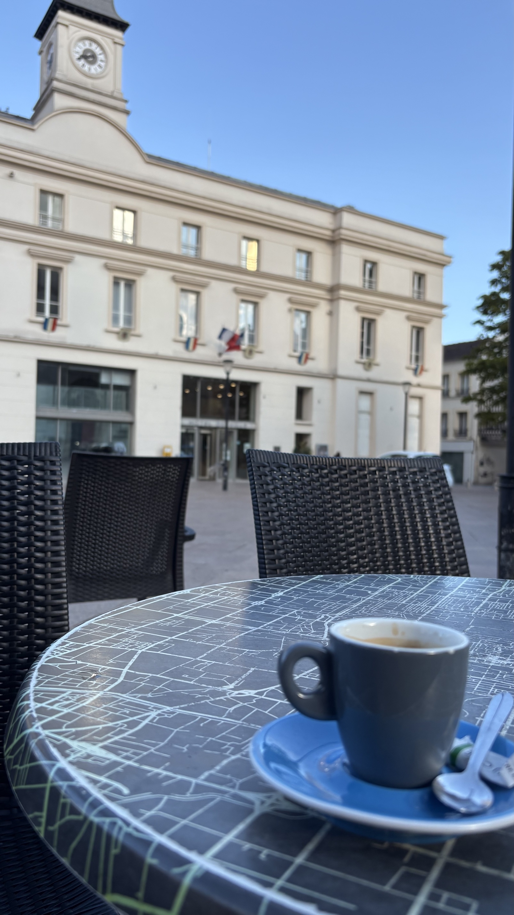 A cup of coffee sits on a patterned table outside near a grand building with a clock tower.