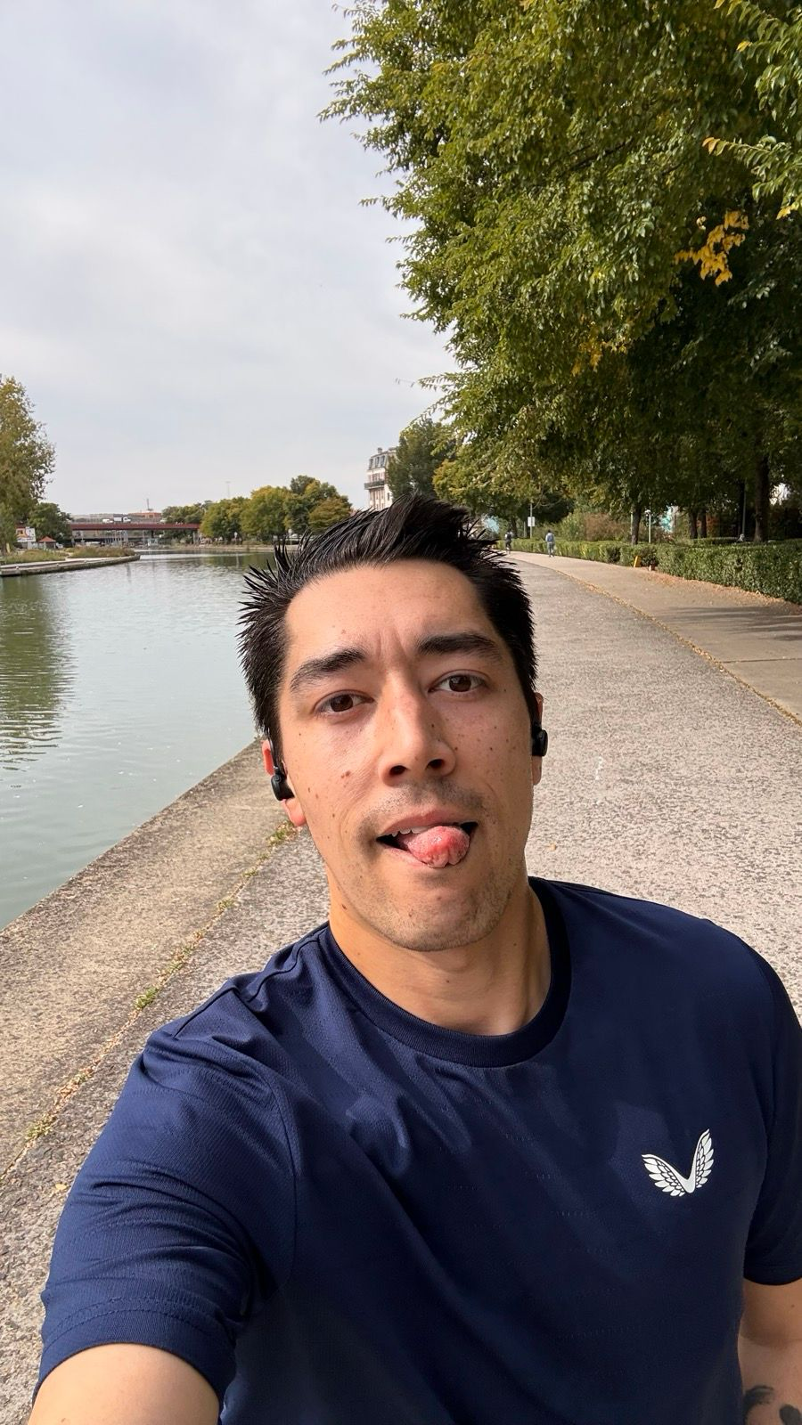 Auto-generated description: A person wearing a blue shirt with earbuds is jogging on a path by a canal lined with trees.