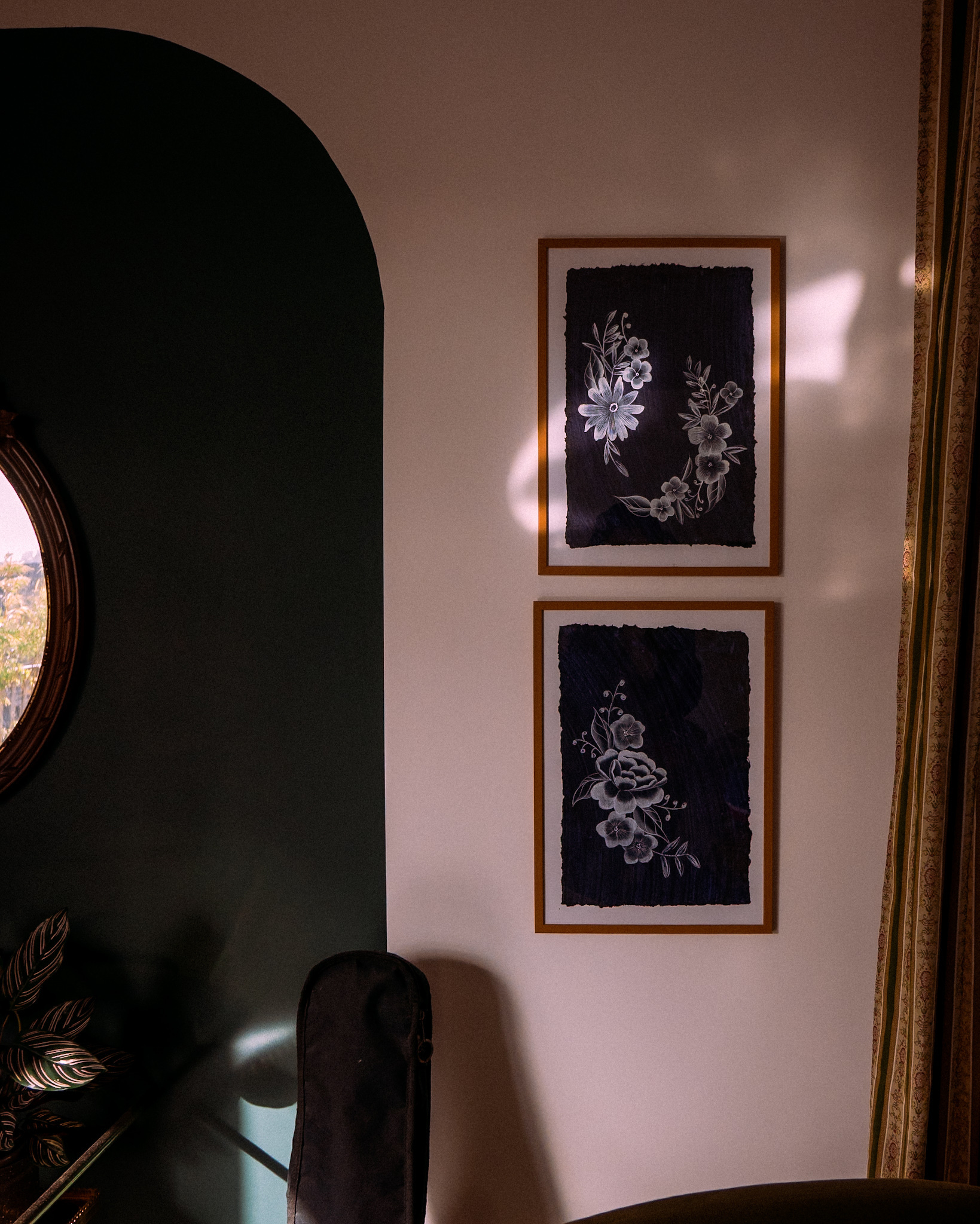 Two framed floral artworks are displayed on a wall with a curved dark green section in a softly lit room.
