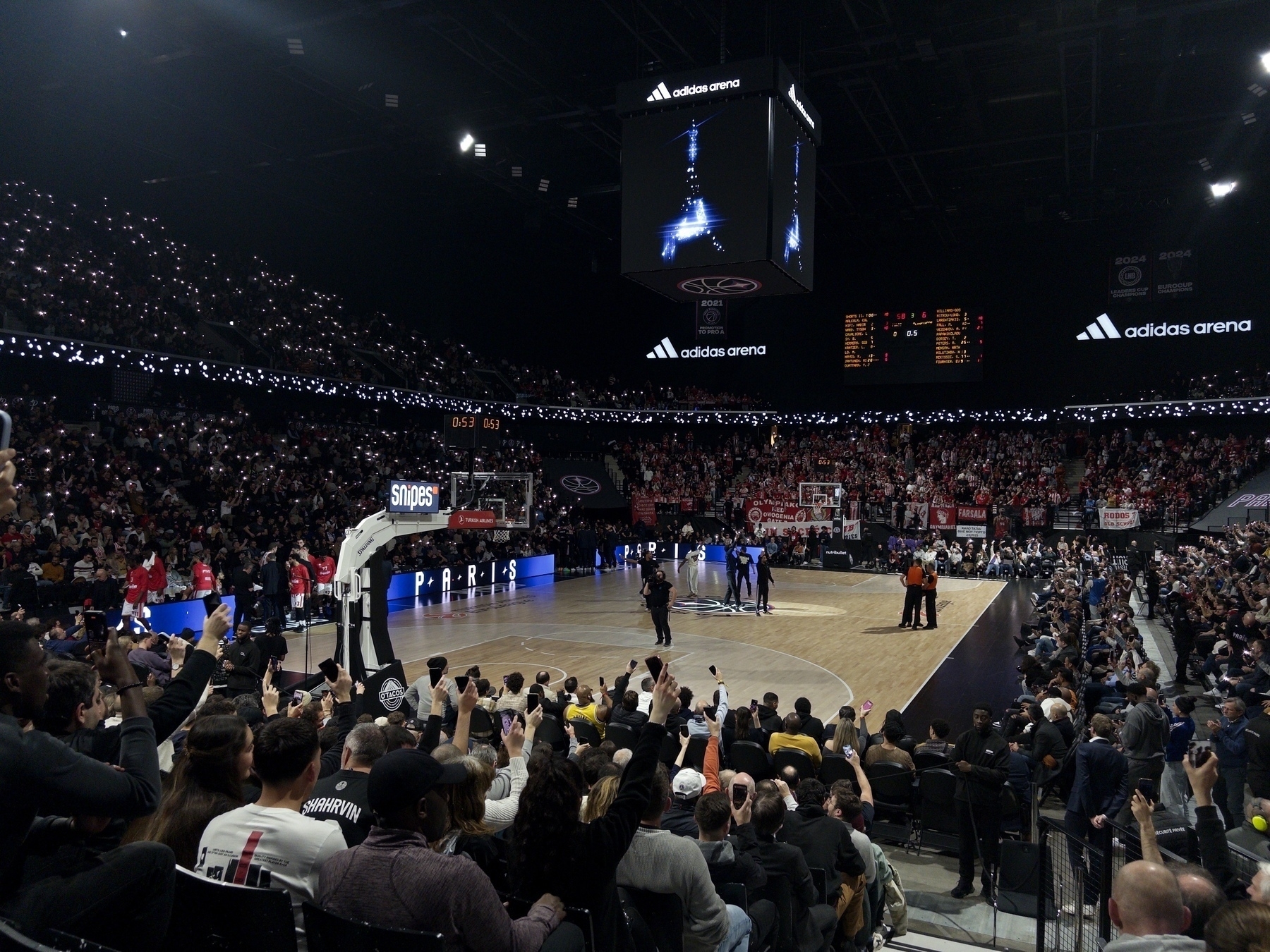 A crowded basketball arena with a large audience is watching a game, featuring players on the court and a scoreboard overhead.