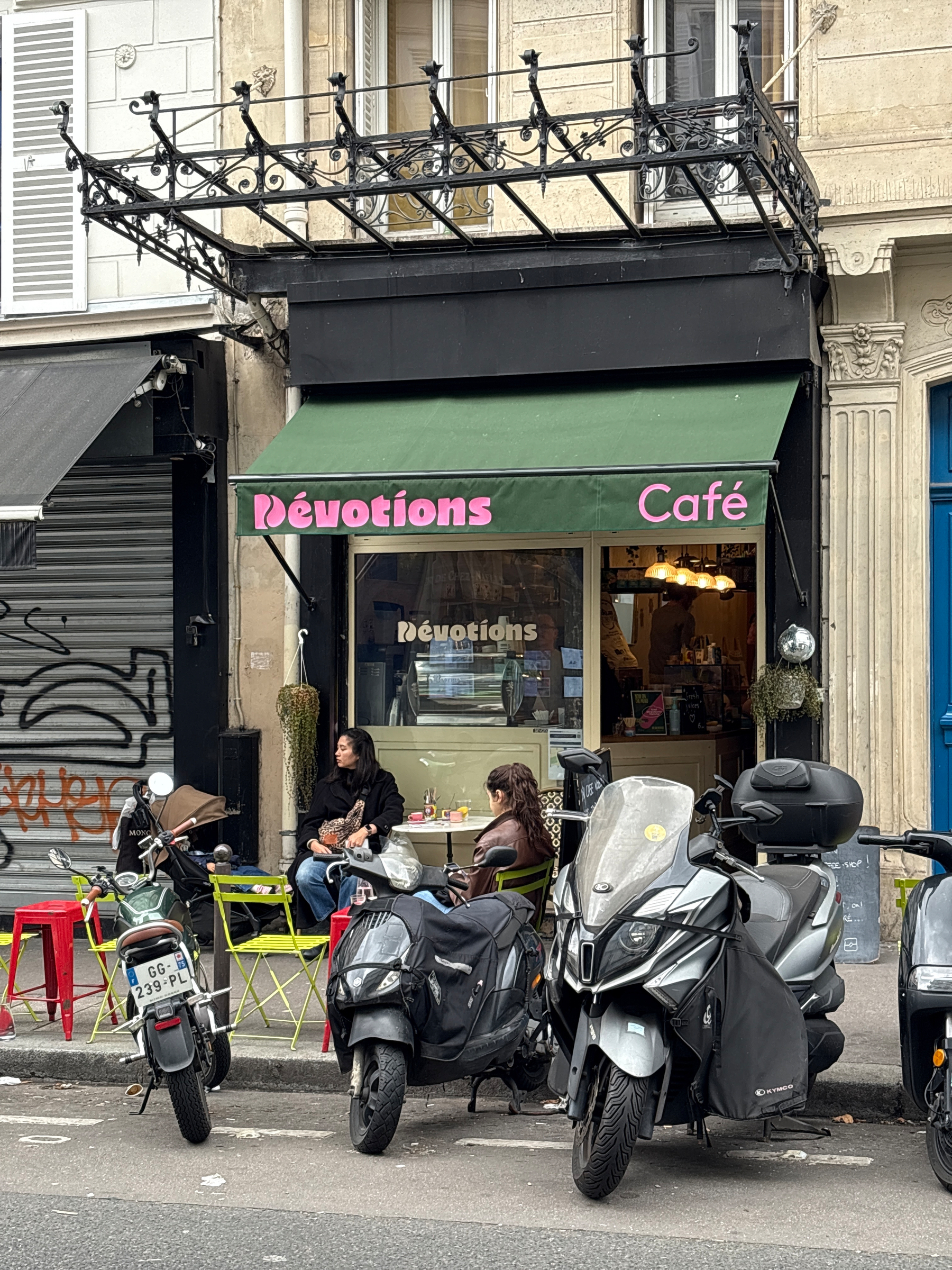 People are sitting outside a café called Dévotions, with scooters parked nearby.