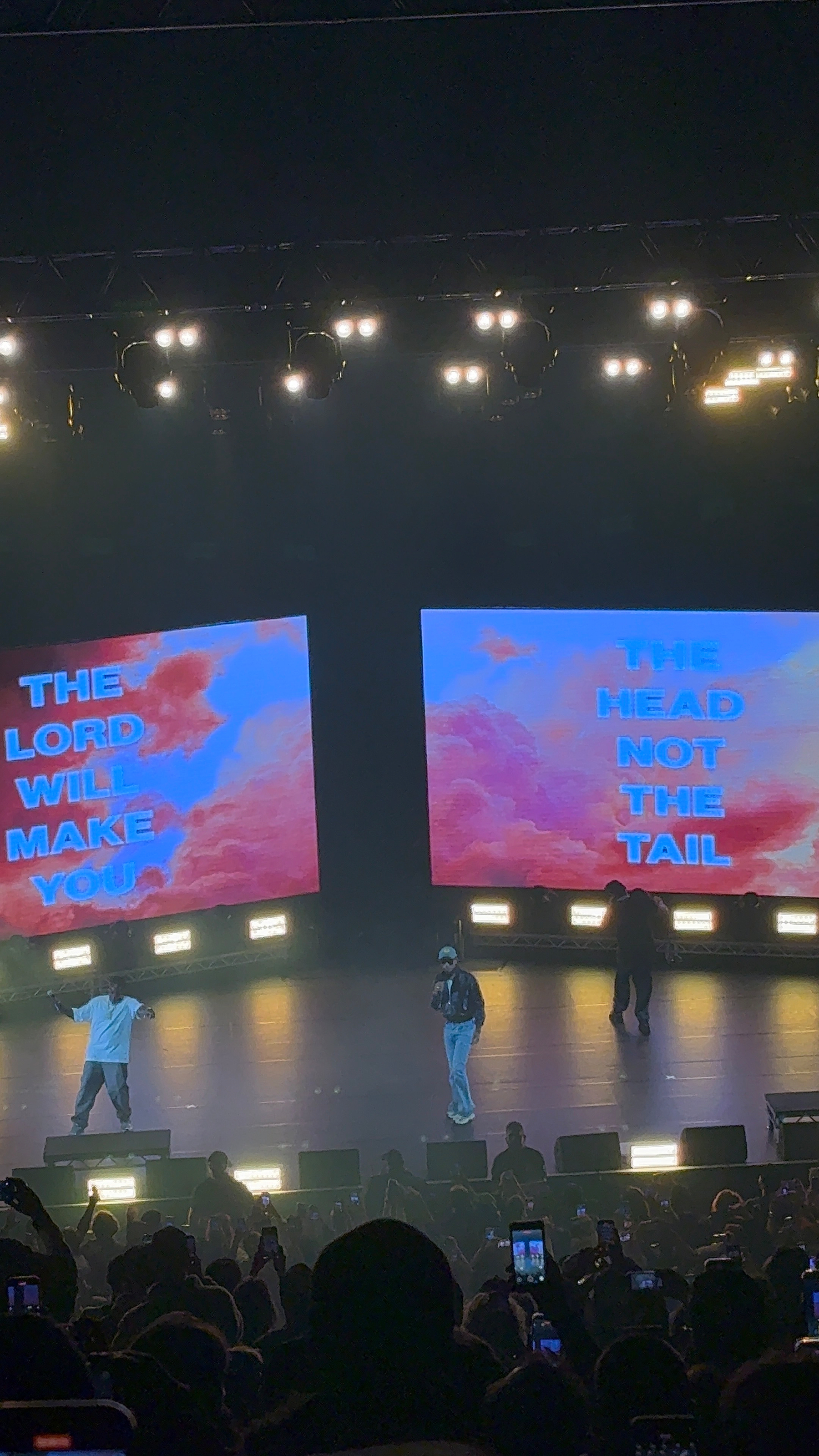 A concert scene with a band performing on stage in front of an audience holding up phones, capturing the moment.