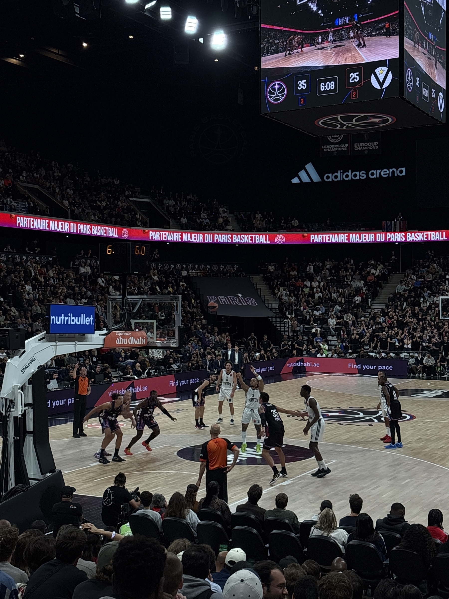 A basketball game is taking place in an indoor arena with a scoreboard and audience in the background.