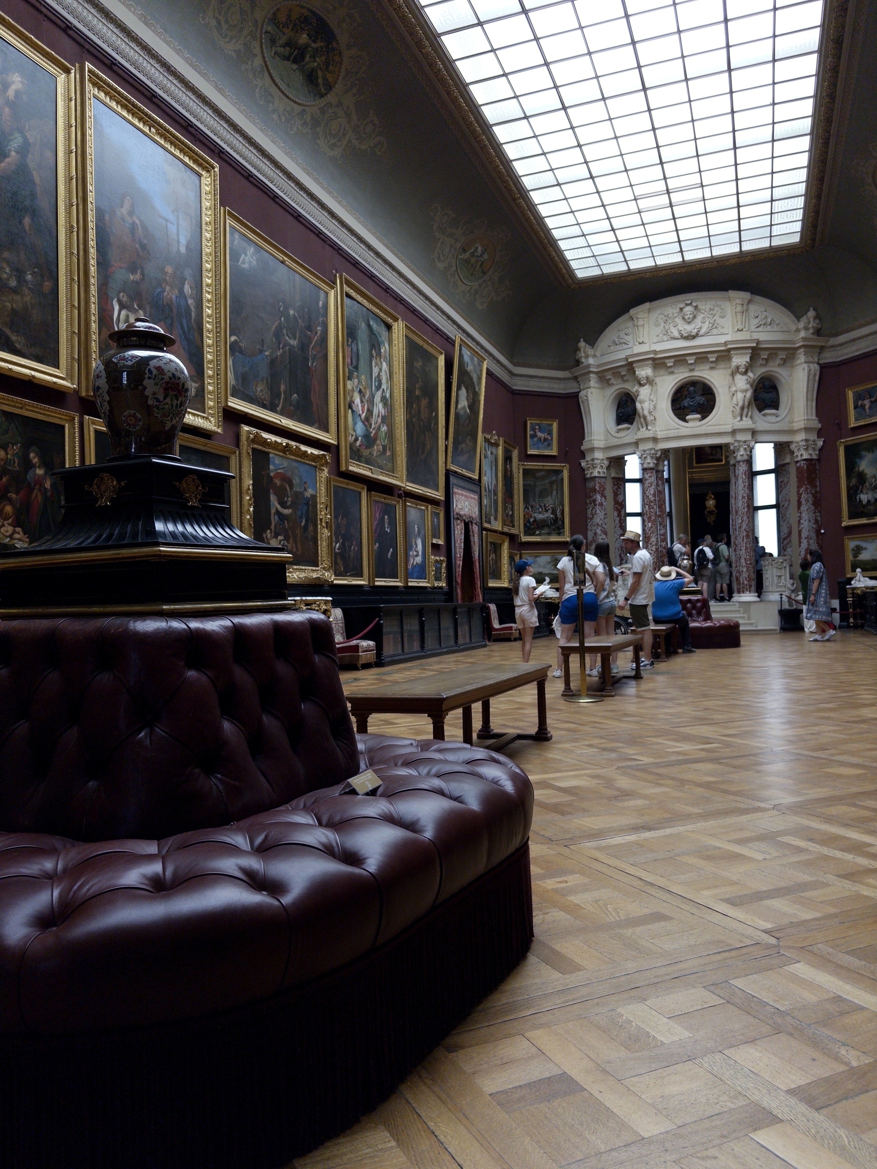 A spacious art gallery features ornate ceiling, wooden floors, and walls adorned with large framed paintings, accompanied by plush seating for visitors.