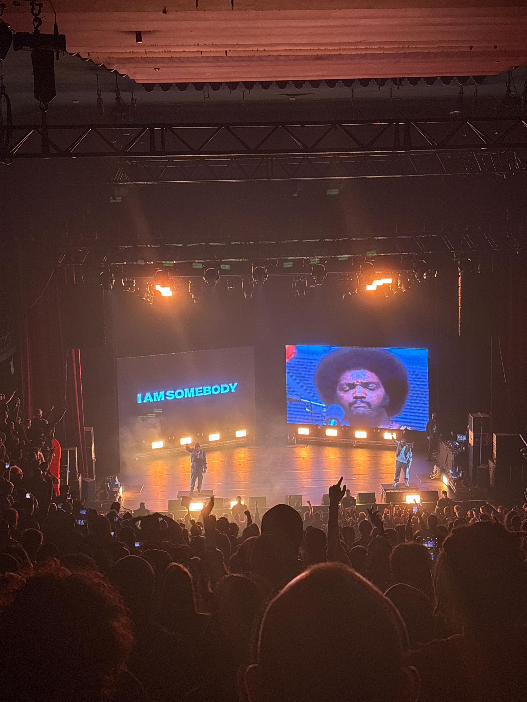 A live performance is taking place on stage with large screens displaying video footage, while the audience captures the scene on their phones.