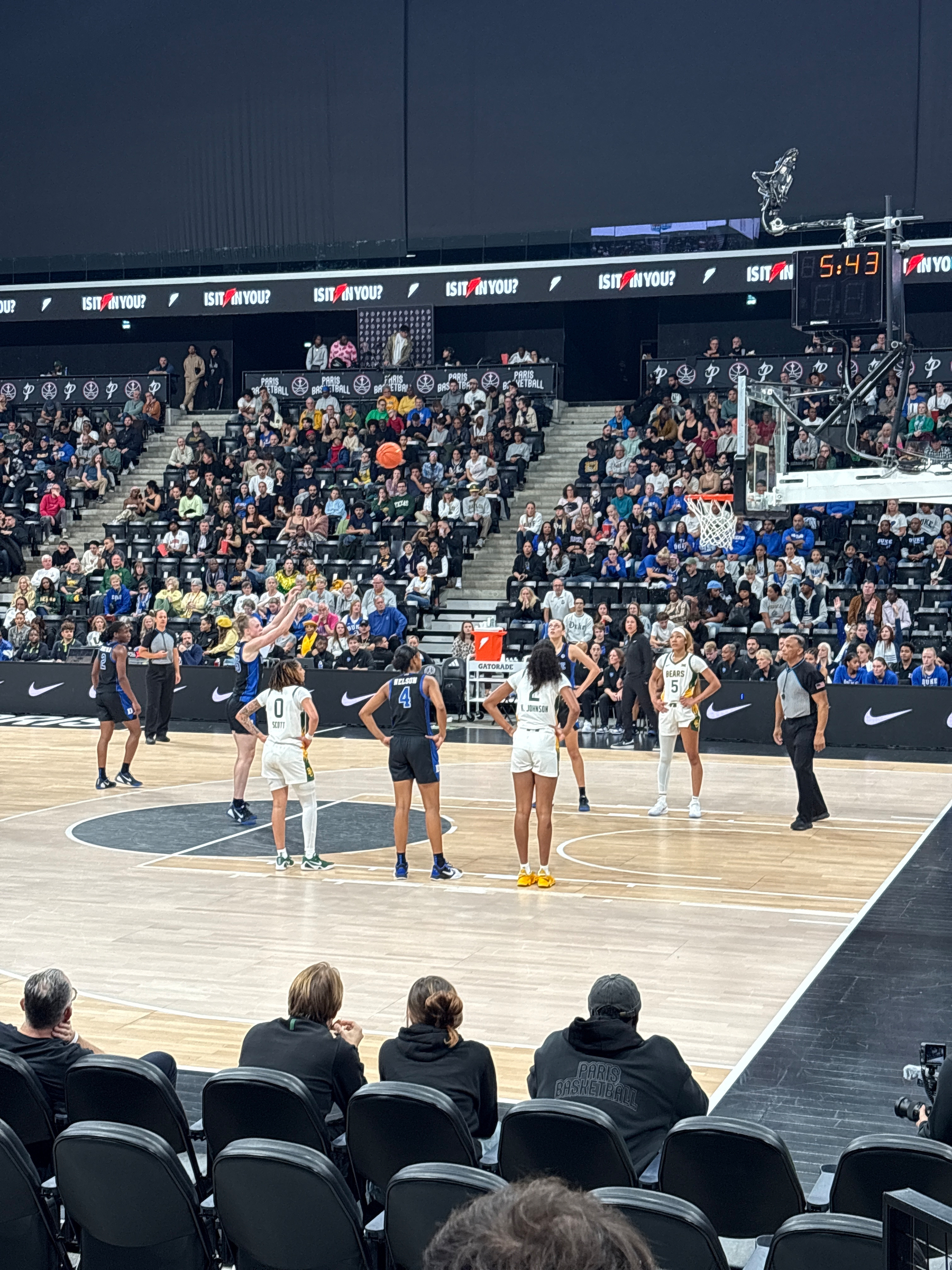 A basketball game is taking place with a player preparing to shoot a free throw while others stand by on the court and spectators watch from the stands.